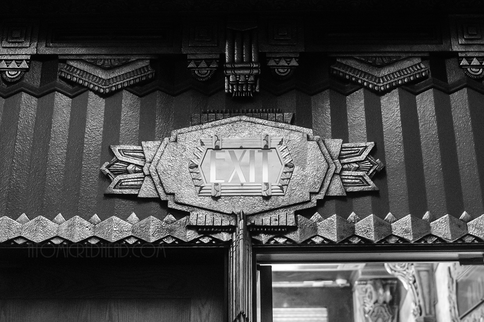 Black and white photo of the exit sign above the doorway, which is framed in an intricate Art Deco design.