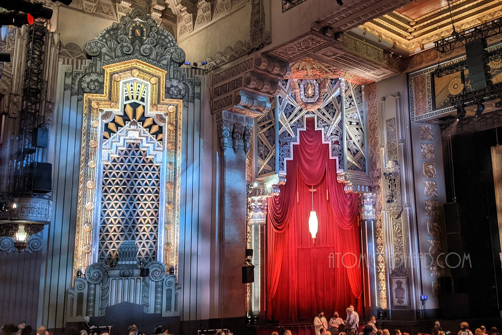 To the right of the stage are two arched openings, one features a red velvet curtain and other has a large grate on it. Art Deco motifs surround the openings.