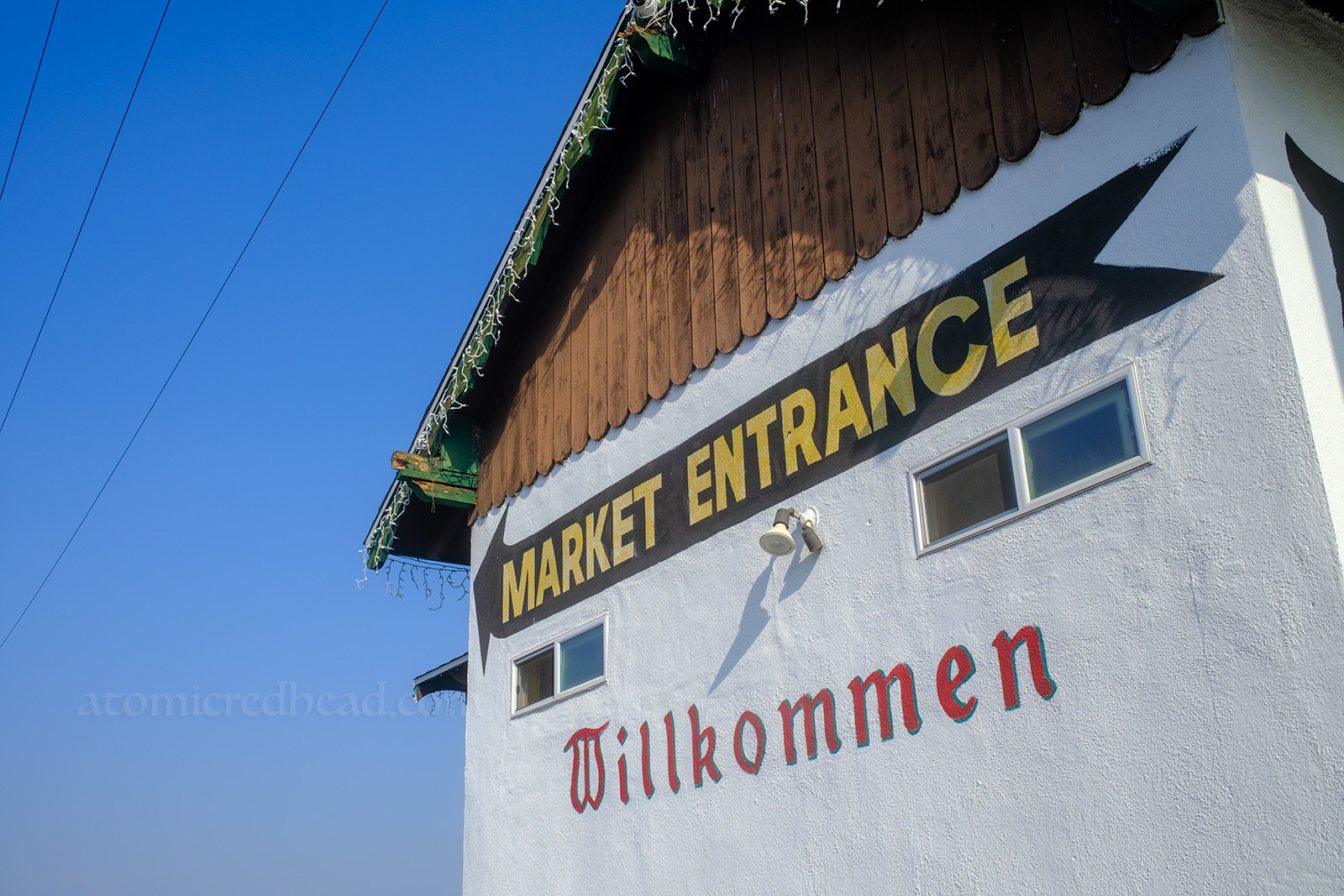 A side of a building has a massive black arrow painted on it, with yellow text reading "Market Entrance" and below, in old fashioned letters reads "Willkommen" meaning "Welcome" in German.