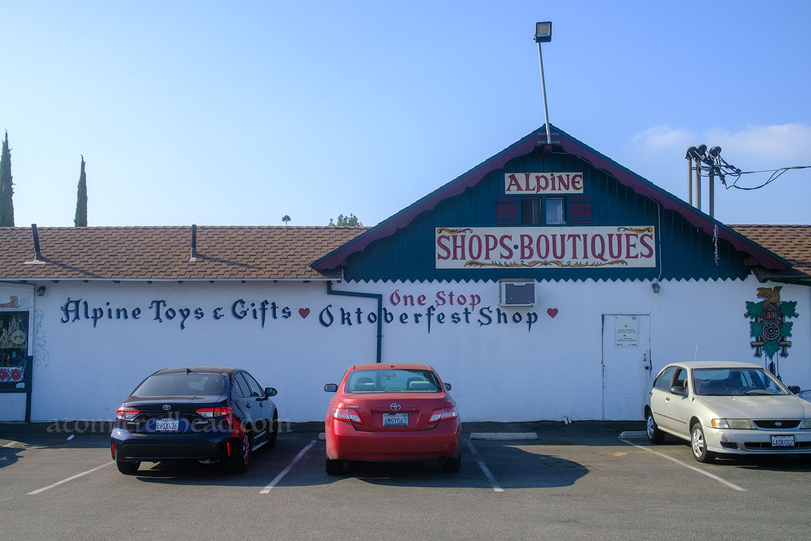 A ski chalet style building features painted signage reading "Alpine Shops Boutiques One Stop Alpine Toys & Gifts Oktoberfest Shop"