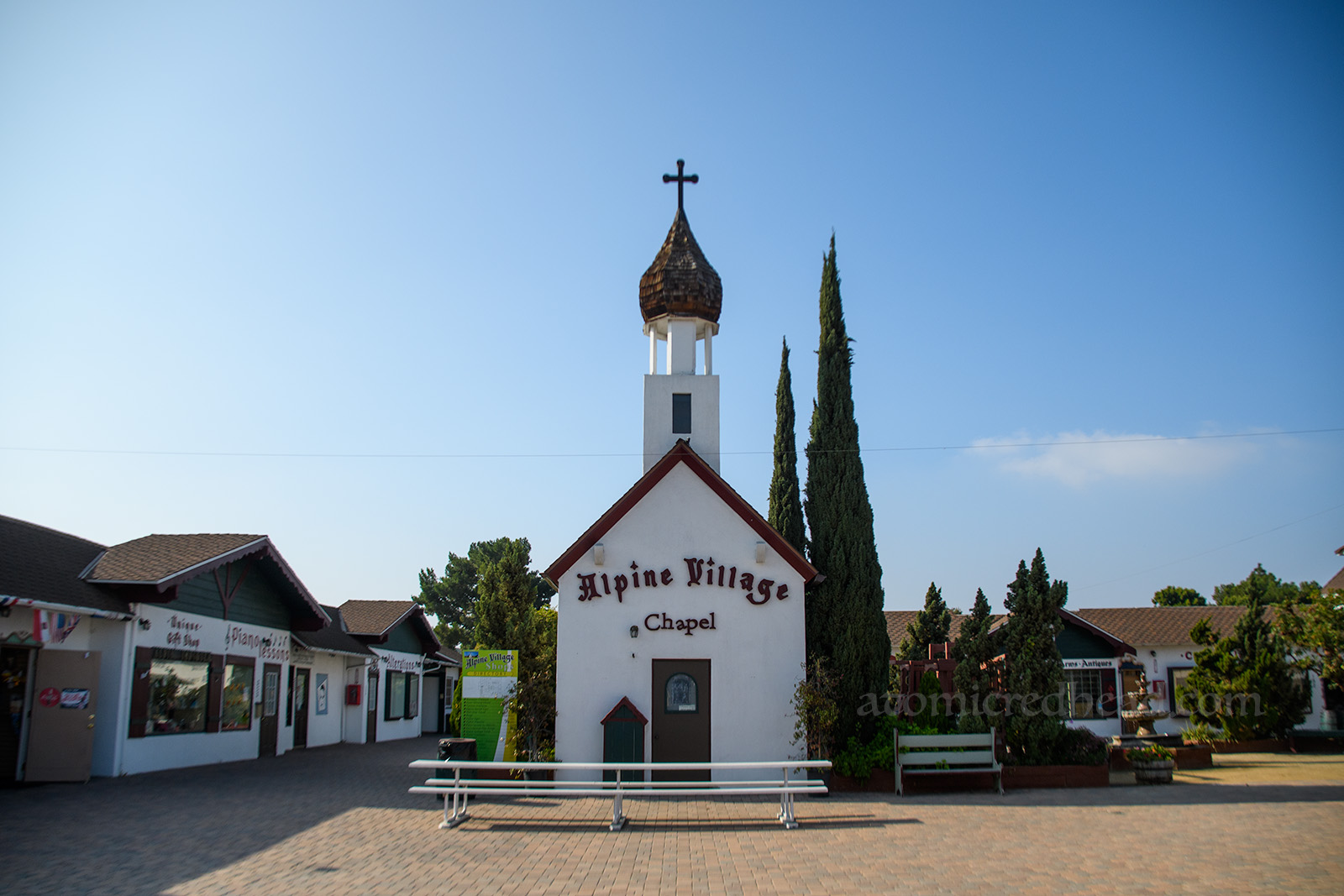 A very small chapel with a spire topped with an onion shaped spire stands in the middle of the small faux German village. Text across the building reads" Alpine Village Chapel."