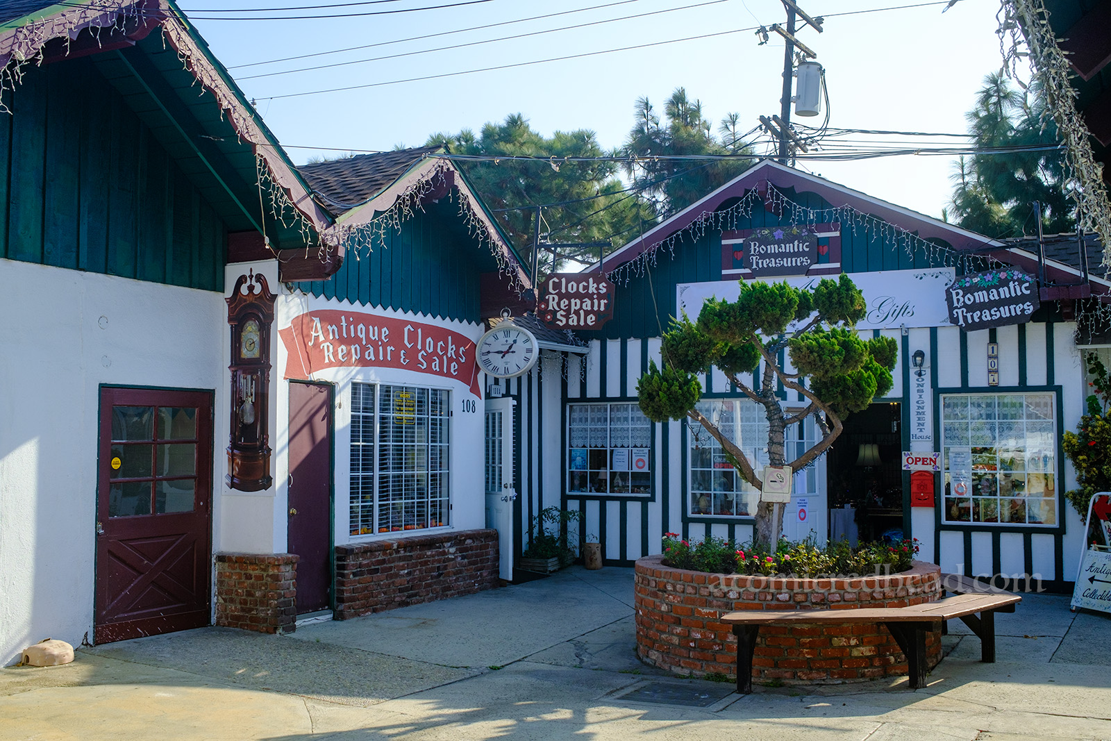 A small corner features two shops, a clock repair and gift shop. The clock repair has a small mural of a grandfather clock, a painted banner reading "Alpine Clocks Repair & Sale" and features a hanging clock in the style of a pocket watch. The gift shop next to it has a small hand painted sign reading "Romantic Treasures." A small brick planter sits in front with a tree inside.