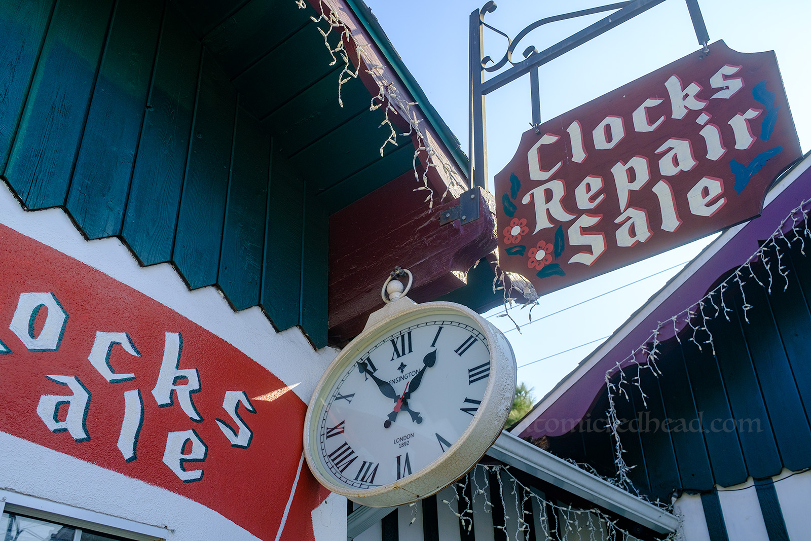 Close-up of the large pocket watch style clock that hangs outside Alpine Clocks. Next to it is a hand painted sign reading "Clocks Repair Sale"