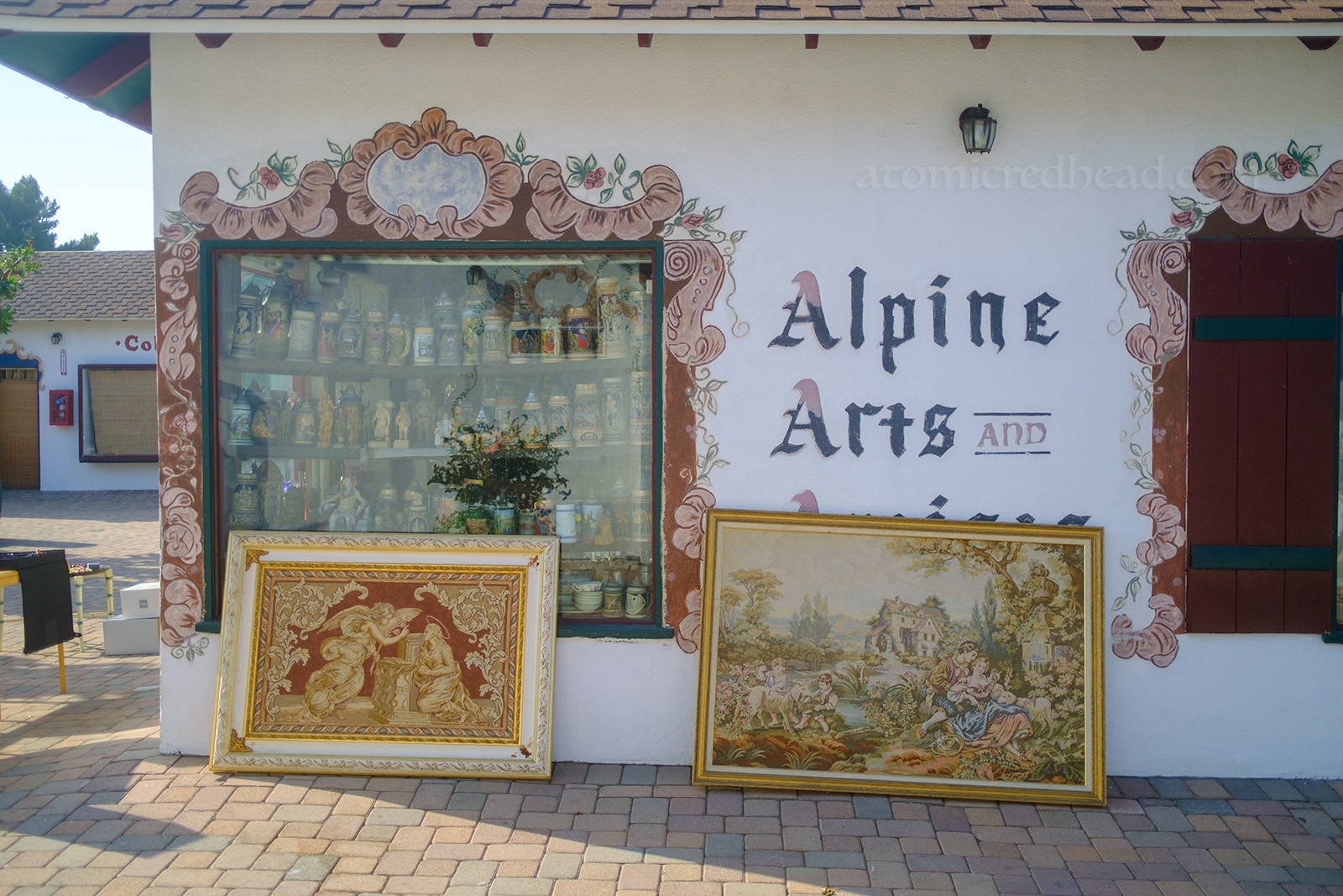 A fanciful frame is painted around the window of a shop. Also painted on the wall reads "Alpine Arts" and large framed works of art sit on the ground, leaning against the building.