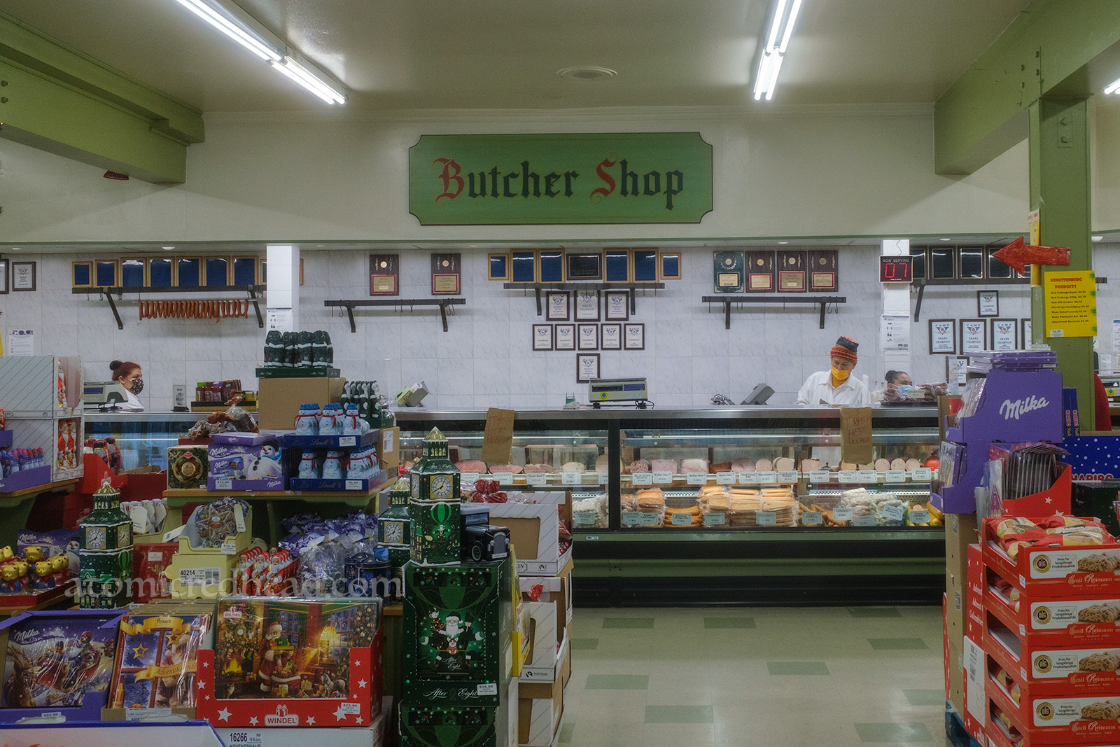 Overall view of part of the market, display cases show off various meats and cheese, a large green sign, hand painted to read "Butcher Shop"