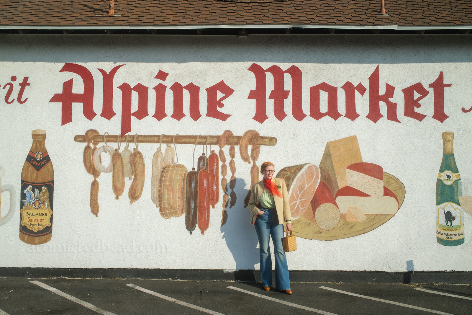 Myself, standing in front of a massive mural of hanging meats and wedges of cheese. Large gothic red letters reads "Alpine Market."
