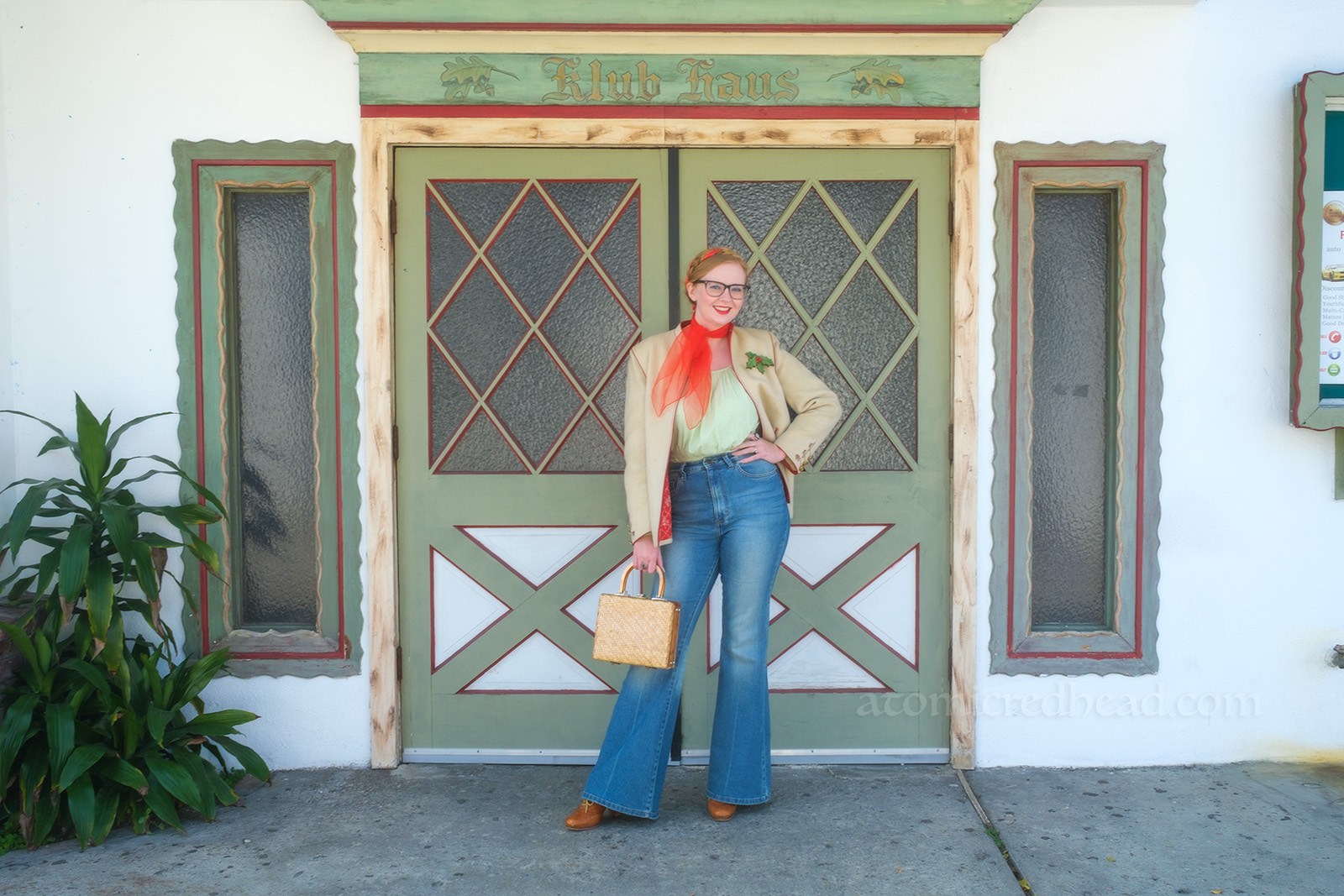 Myself, wearing a white jacket, red scarf around my neck, light green peasant blouse, and blue jeans, standing in front of a white and green set of double doors. Above rads "Klub Haus"