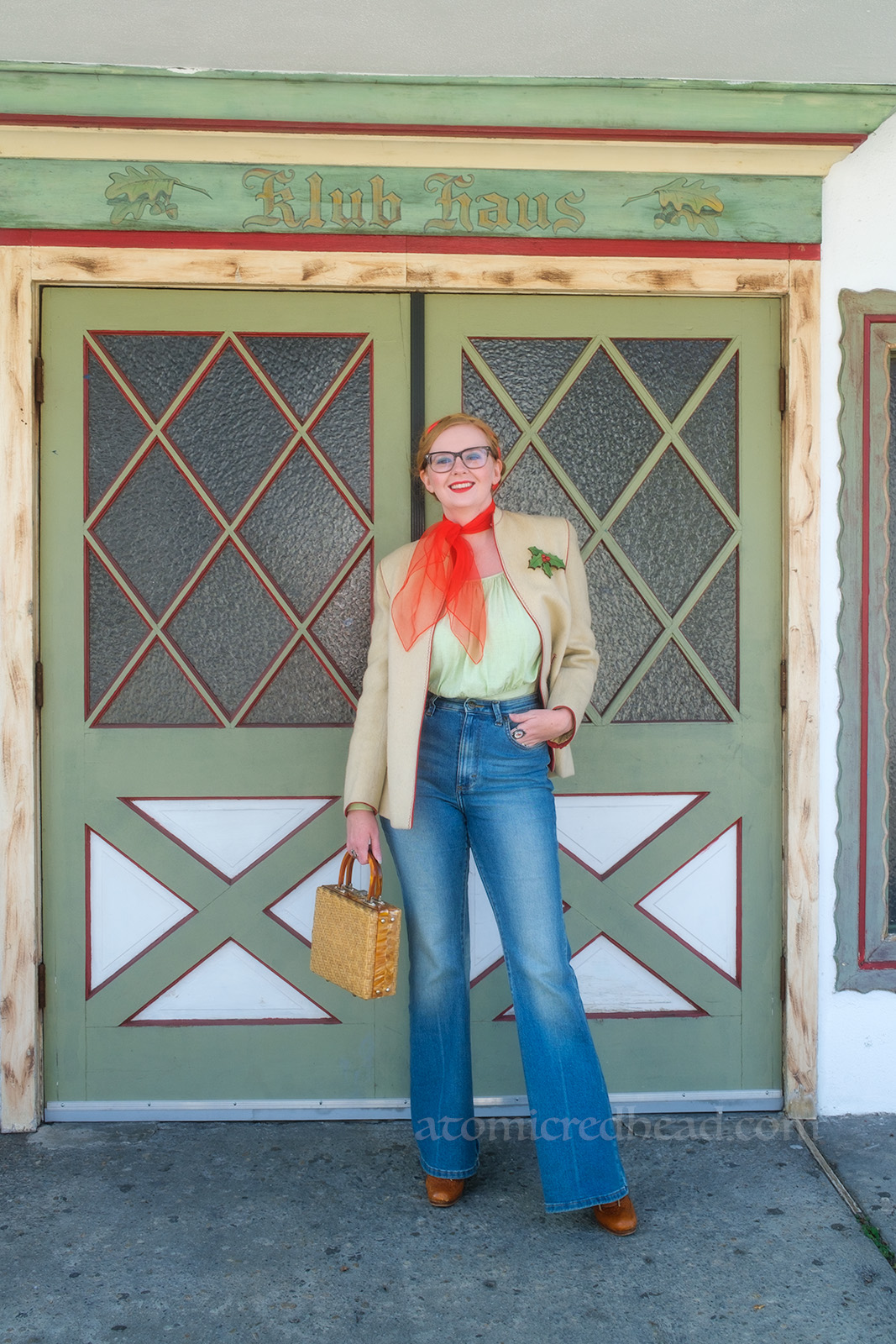Myself, wearing a white jacket, red scarf around my neck, light green peasant blouse, and blue jeans, standing in front of a white and green set of double doors. Above rads "Klub Haus"