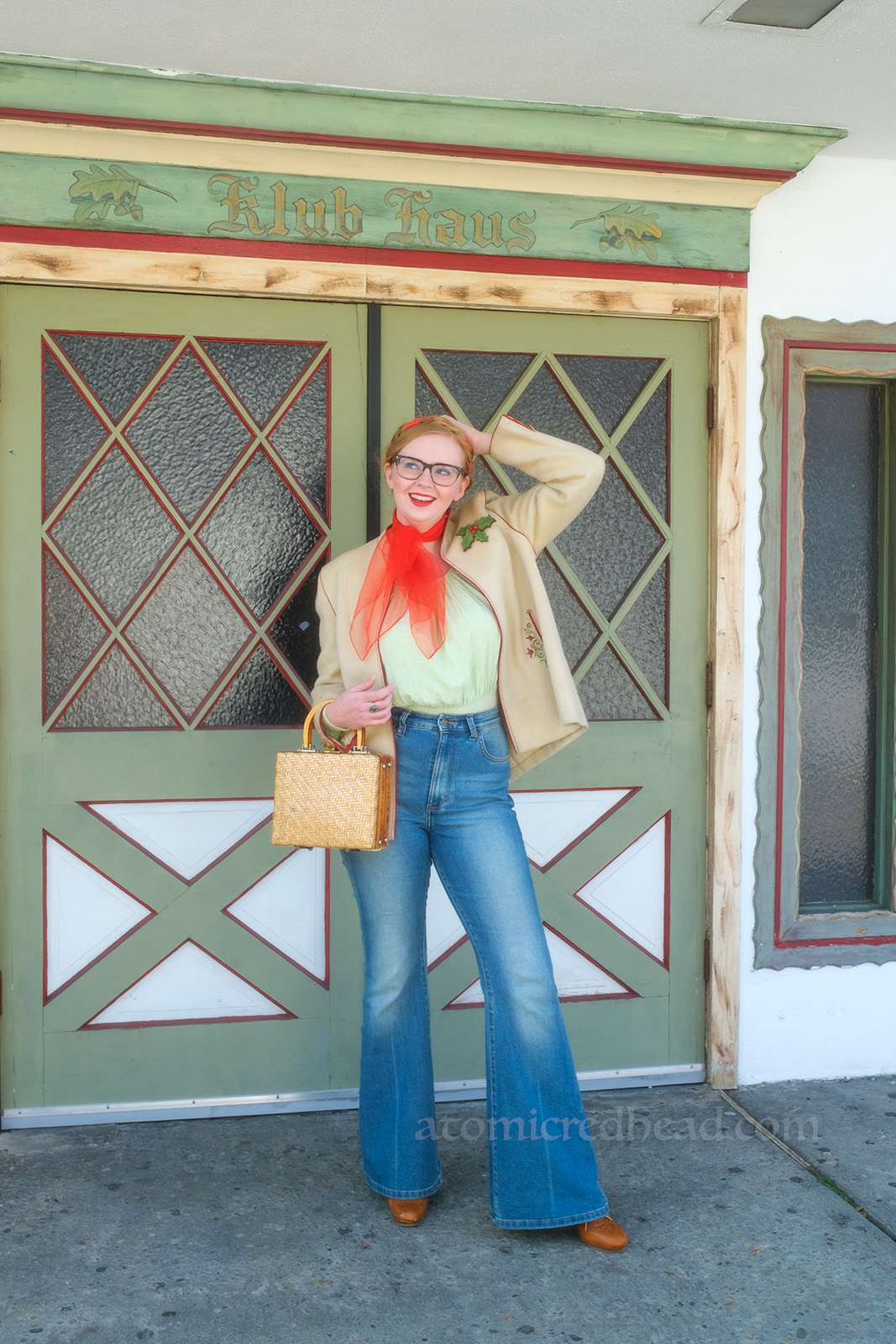 Myself, wearing a white jacket, red scarf around my neck, light green peasant blouse, and blue jeans, standing in front of a white and green set of double doors. Above rads "Klub Haus"