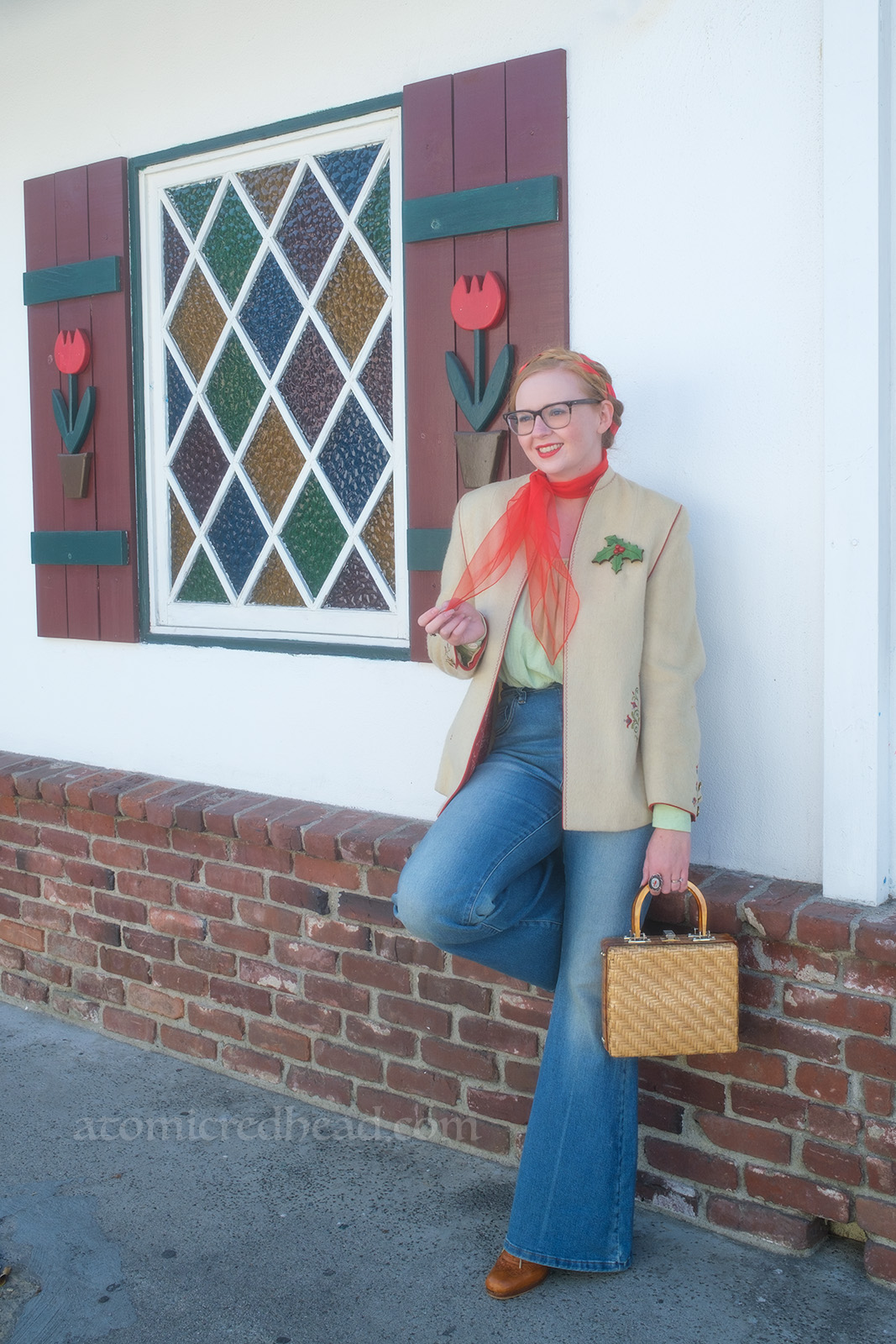 Myself, wearing a white jacket, red scarf around my neck, light green peasant blouse, and blue jeans, leaning against a wall, behind me a stained glass diamond pane window also features shutters with tulips on them.