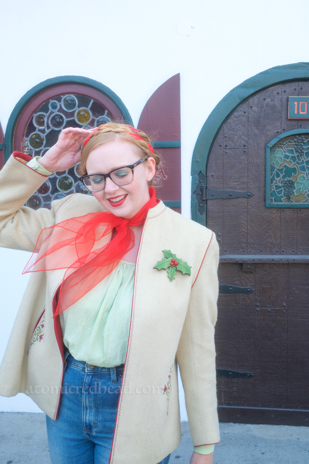 Myself, wearing a white jacket, red scarf around my neck, light green peasant blouse, and blue jeans, standing in front of a white and green set of double doors. Behind me an arched door features stained glass of grapes.