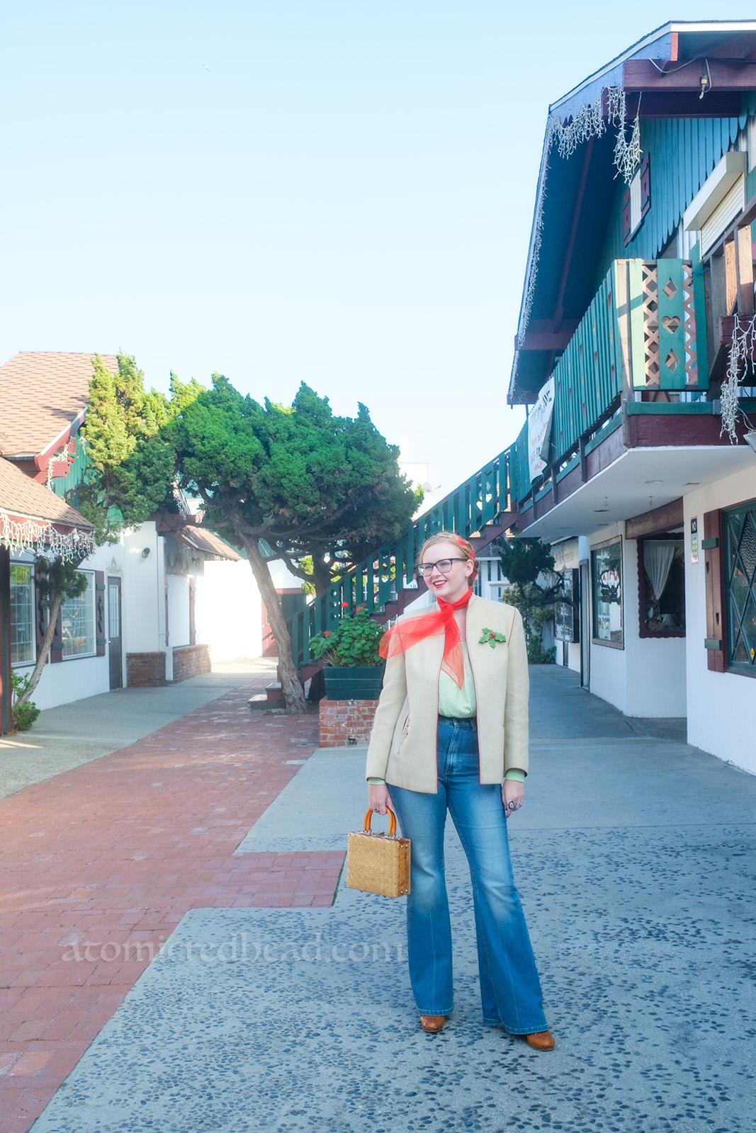Myself, wearing a white jacket, red scarf around my neck, light green peasant blouse, and blue jeans, standing in the middle of one of the walkways of the village.