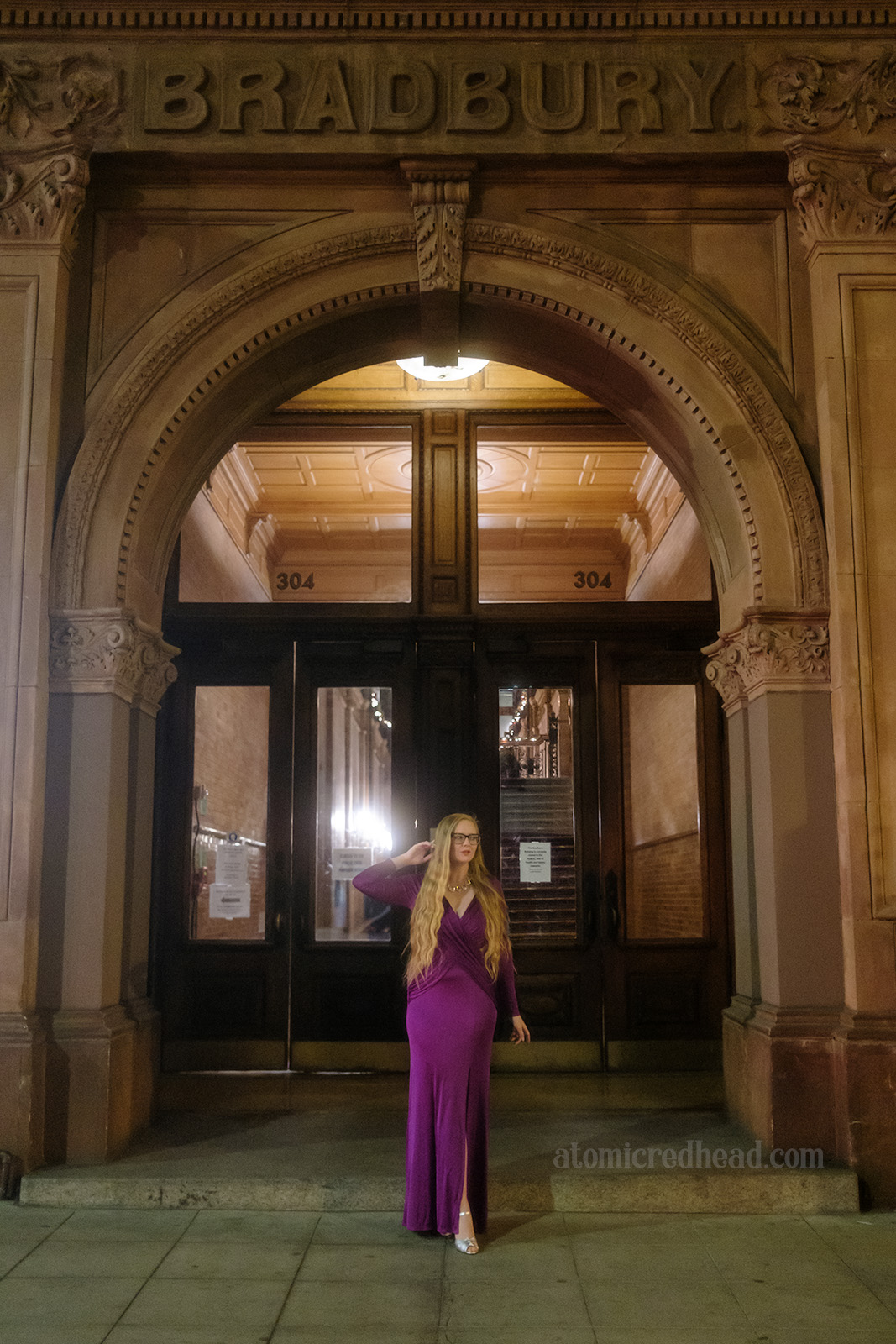 Myself, wearing a full length purple dress, standing outside the Bradbury Building, a brick building with an arched entryway with dark doors leading inside.