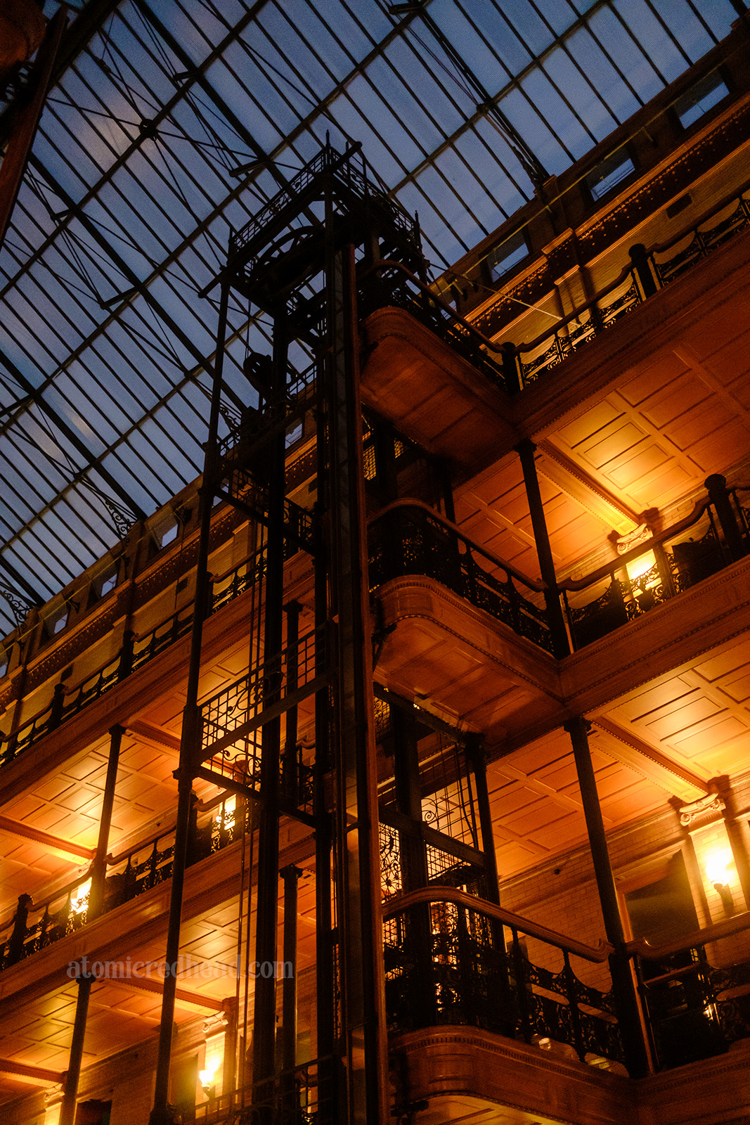A black, wrought iron cage style elevator shaft reaches toward a central skylight. Open balconies flank the elevator with warm wood walls and ceilings.