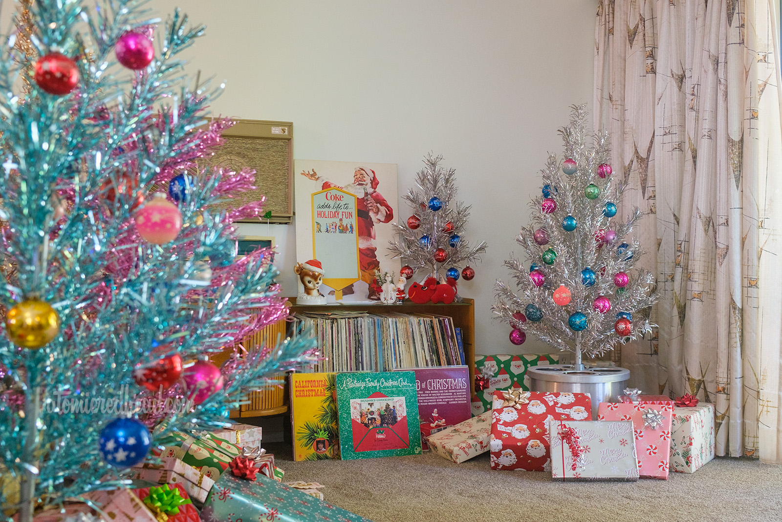 In the foreground on the left, a blue aluminum tree peeks into frame on the right a 4 foot tall silver aluminum tree sits. Both have colorful ball ornaments on them.