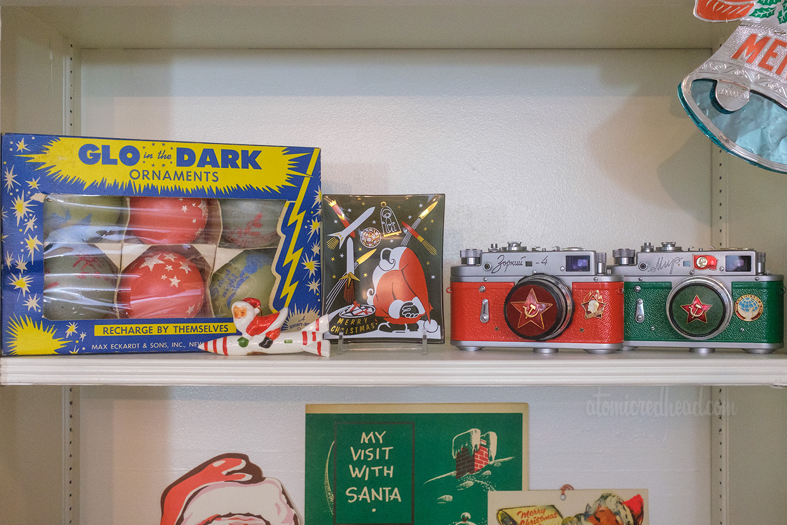 "Glow in the Dark" Christmas ornaments sit on a shelf, in their original box, with stars on them, a small glass dish features Santa looking to the sky which is filled with rockets, a small ceramic ornament of Santa on a rocket sits in front, and to the right sit two cameras, one red, one green, that are from the Soviet Union.