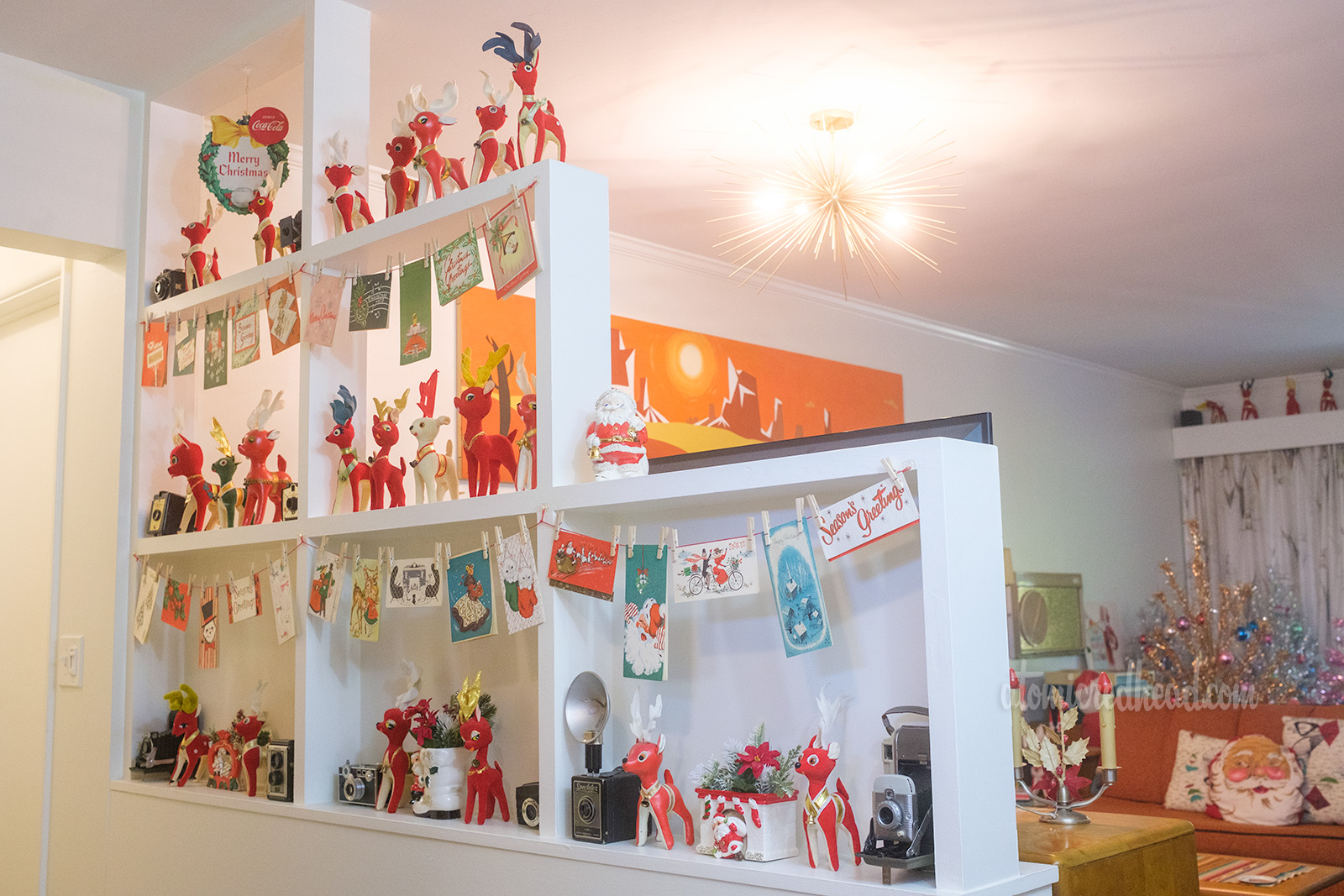 A build in room divider separates the living room from the hallway. It features various Christmas cards, felt reindeer and several ceramic planters featuring Santa filled with greenery.