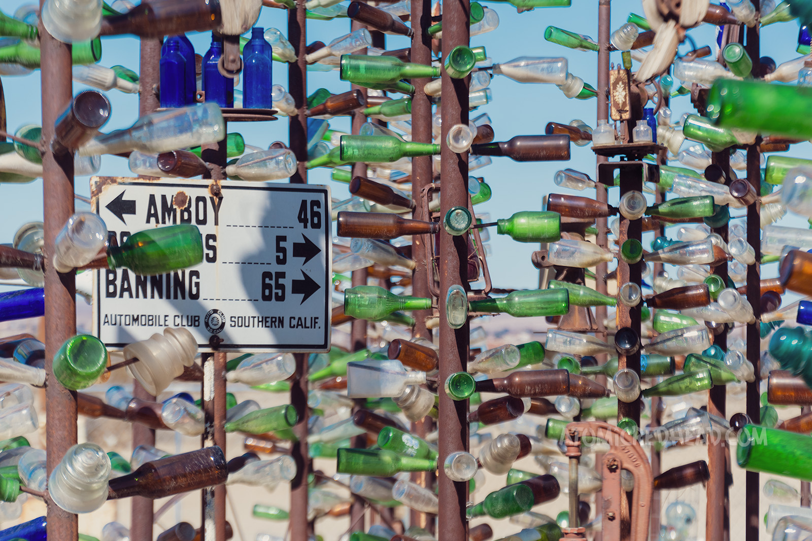 Somewhat wide shot showcasing the middle section of various "bottle trees" which are tall metal posts with smaller rods jutting out from the side, with glass bottles slid onto them.
