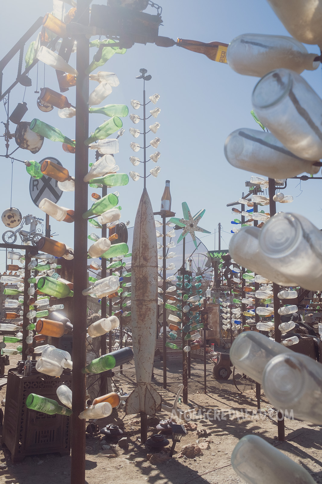 Wide, overall shot of Bottle Tree Ranch. The "bottle trees" are tall metal posts with smaller rods jutting out from the side, with glass bottles slid onto them. Some of the trees are topped with various items, such as wheels, lanterns, and signs. In the middle a missile stands on its end.