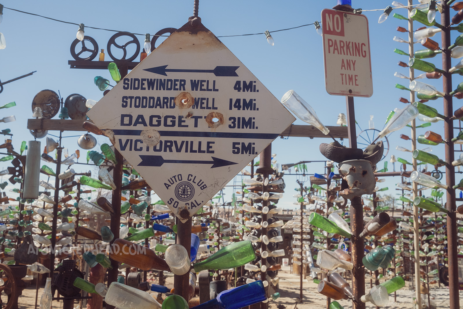 One tree is topped with a sign reading the distances to other desert cities, such as Sidewinder Well, Stoddard Well, Dagget, and Victorville.