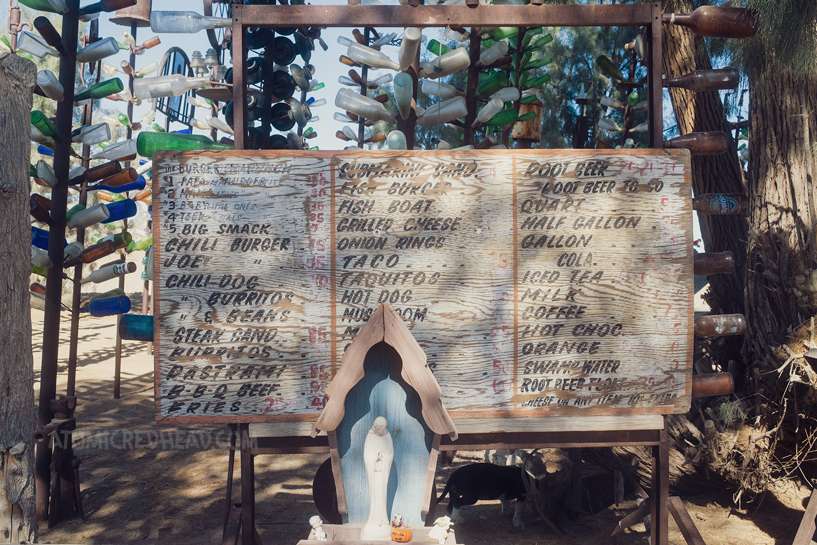 A vintage, hand painted menu board, featuring items such as burgers, tacos, and beverages, all with prices under $1. A small statue of Mary sits in front, various tall bottle trees are behind the menu.