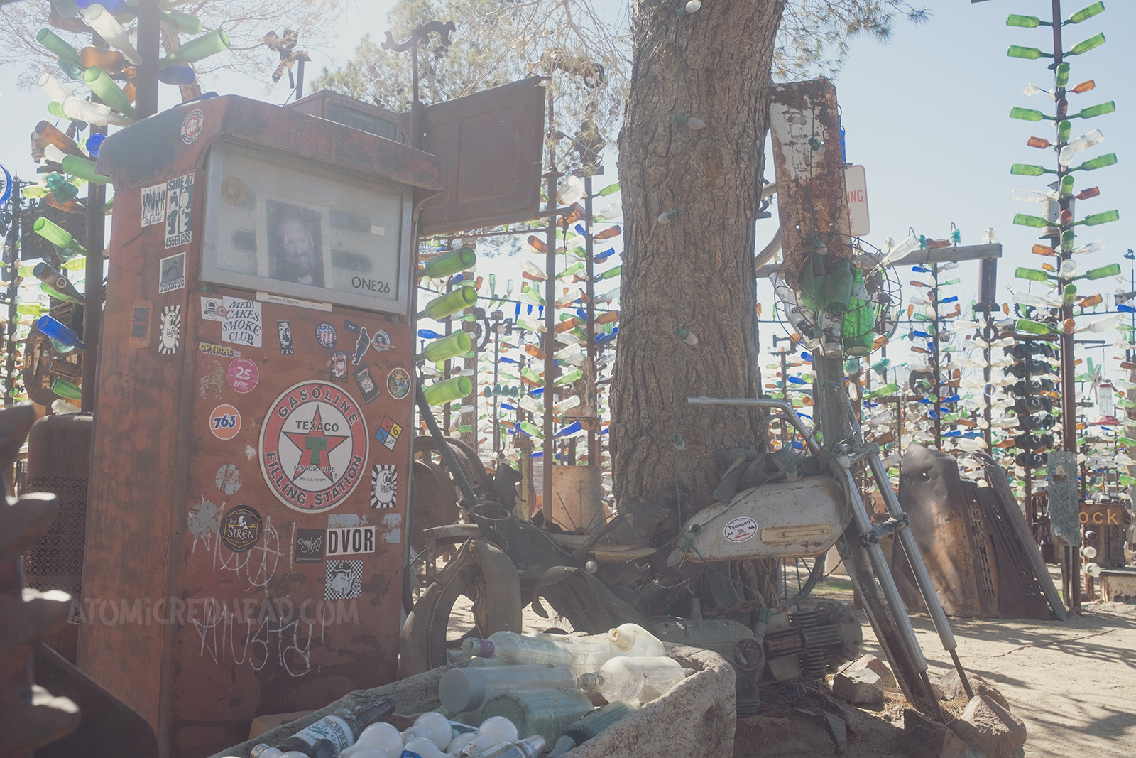 An old gas pump, covered with stickers from visitors, an old motorcycle, and a trough filled with vintage bottles sit under the shade of a real tree.