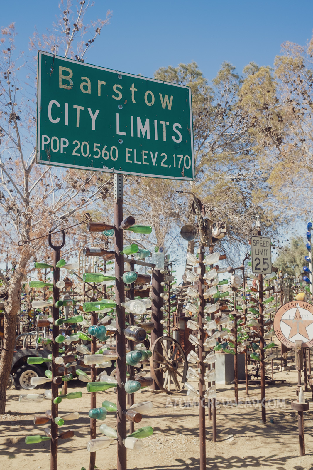 A sign reading "Barstow City Limits Pop. 20,560 Elev. 2170" sits atop a bottle tree.