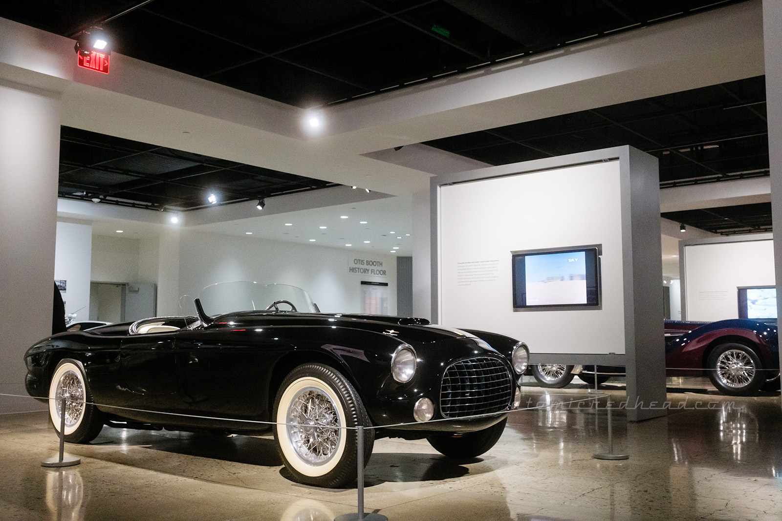 An early 50s black Ferrari sits in a wide open space in the museum, other cars visible in the distace.