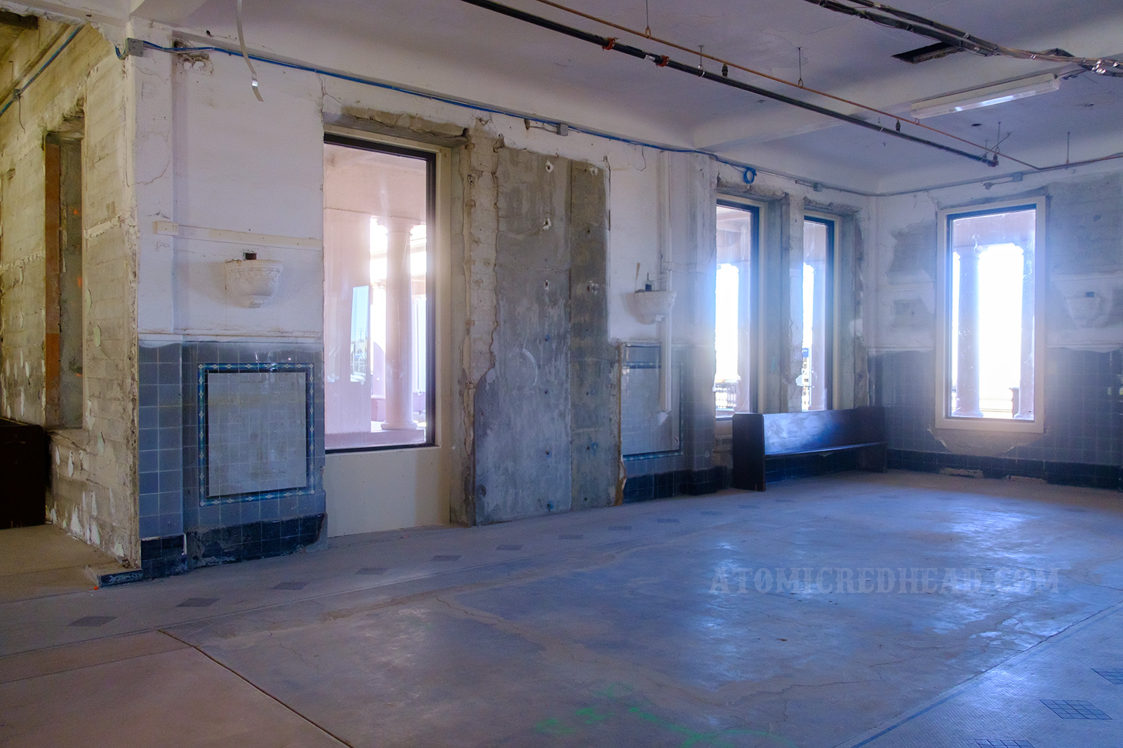 A corner of the former lunch counter. Windows look out toward the tracks, sconces flank the walls, some of the walls feature grey, black, and blue tiles with a small floral design. On the floor a tile design showcases the footprint of the old lunch counter.