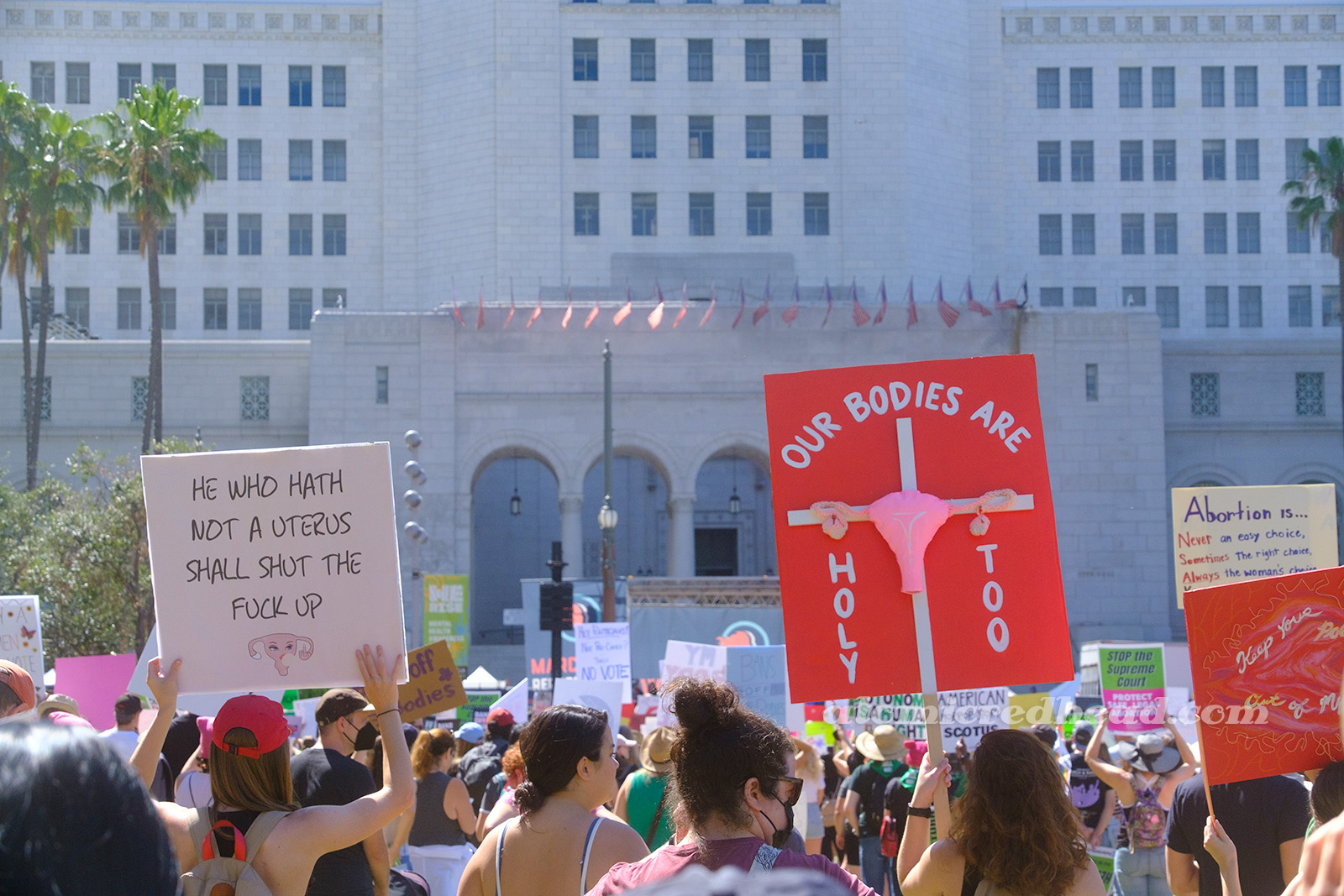 Two signs, one on the left reads "He who hath not a uterus shall shut the fuck up." the other features a uterus on a cross and text reading "Our Bodies are Holy Too"