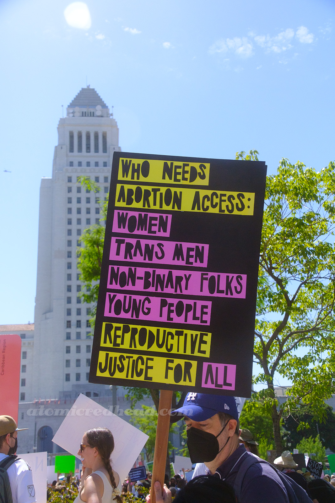 A black sign with pink and yellow letters reads "Who Needs Abortion Access: Women, Trans Men, Non-binary Folks, Young People. Reproductive Justice For All"