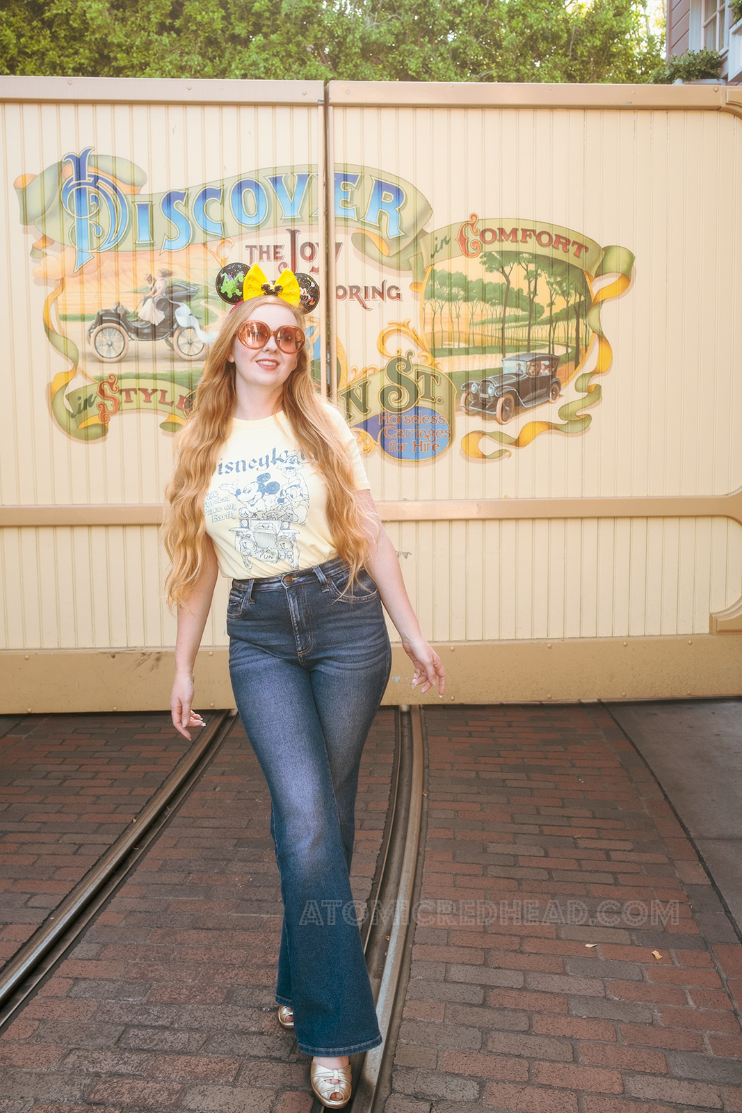 Myself, standing in front of a Main Street mural, wearing special Main Street Electrical Parade Mickey ears, which feature Elliot the green dragon, and Mickey, a butter yellow shirt featuring Mickey, Donald, and Goofy in an old jalopy, with text reading "Disneyland The Happiest Place on Earth"