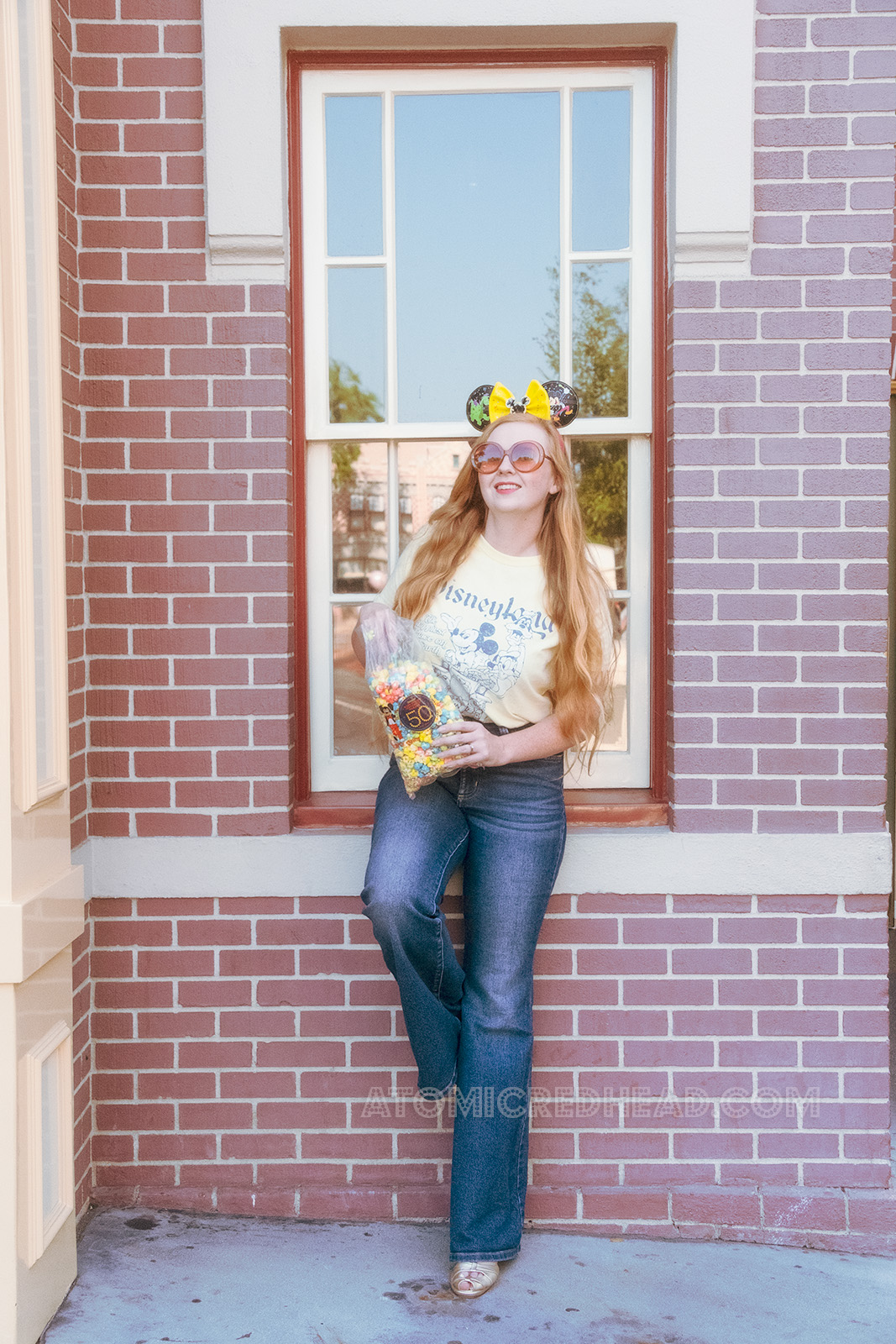 Myself, standing in front of the Disneyland Fire Station, wearing special Main Street Electrical Parade Mickey ears, which feature Elliot the green dragon, and Mickey, a butter yellow shirt featuring Mickey, Donald, and Goofy in an old jalopy, with text reading "Disneyland The Happiest Place on Earth" and holding a bag of colorful kettle corn that has a sticker on it celebrating the 50th anniversary of the Main Street Electrical Parade.