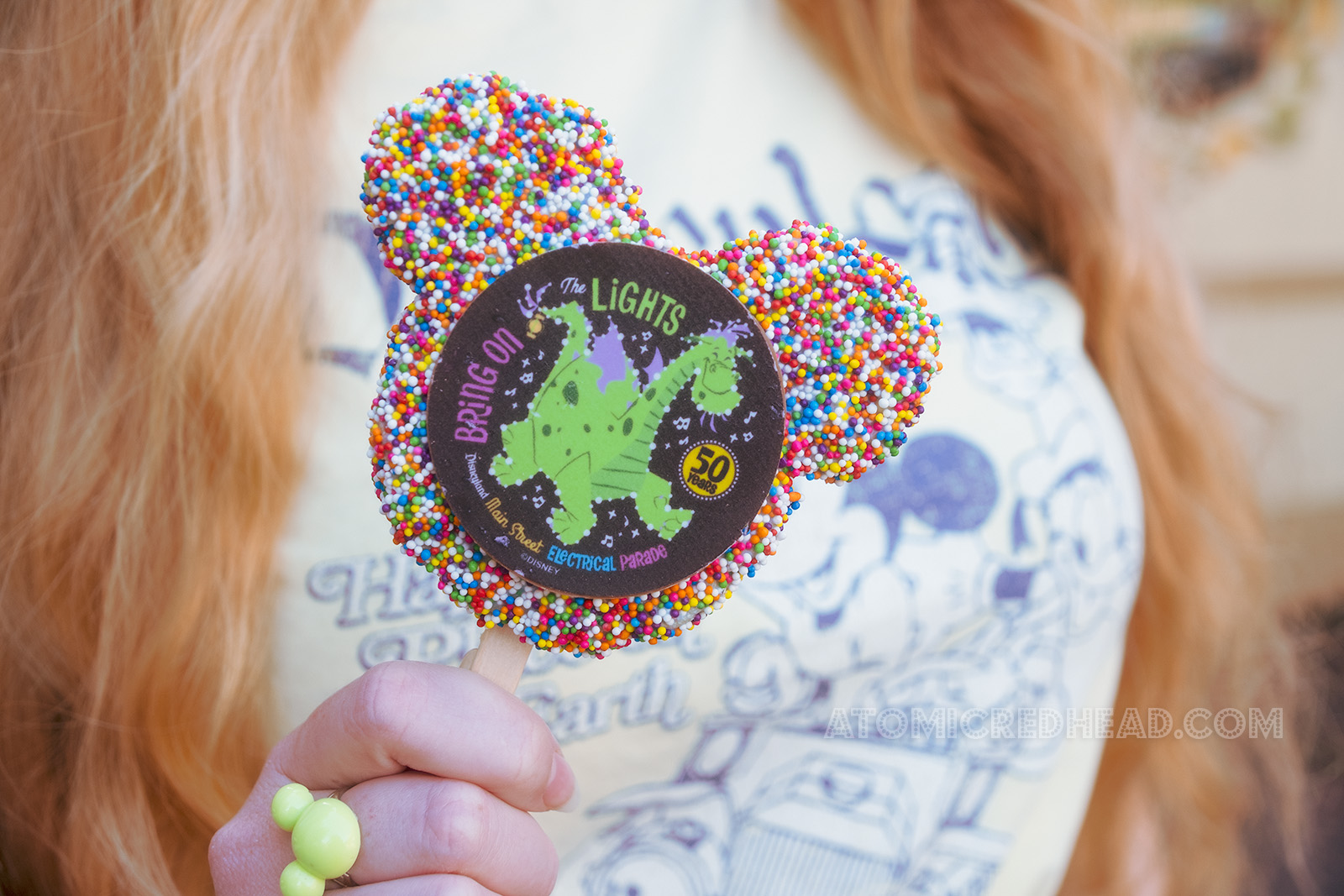 Close-up of a Mickey shaped rice crispy treat pop, covered in colorful sprinkles and a chocolate disk featuring Elliot the green dragon, and text reading "Bring on the Lights! 50 years Disneyland Main Street Electrical Parade"
