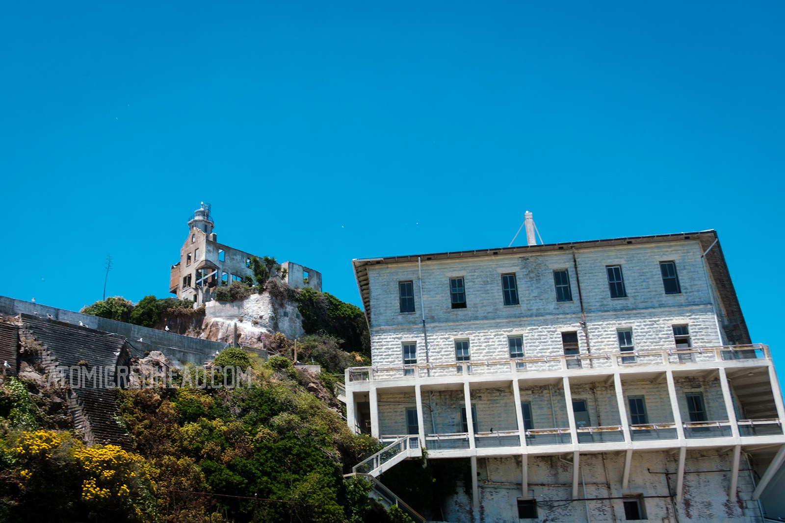 The former barracks, a three story white building juts out from the edge of the island, with the lighthouse visible above and to the left.