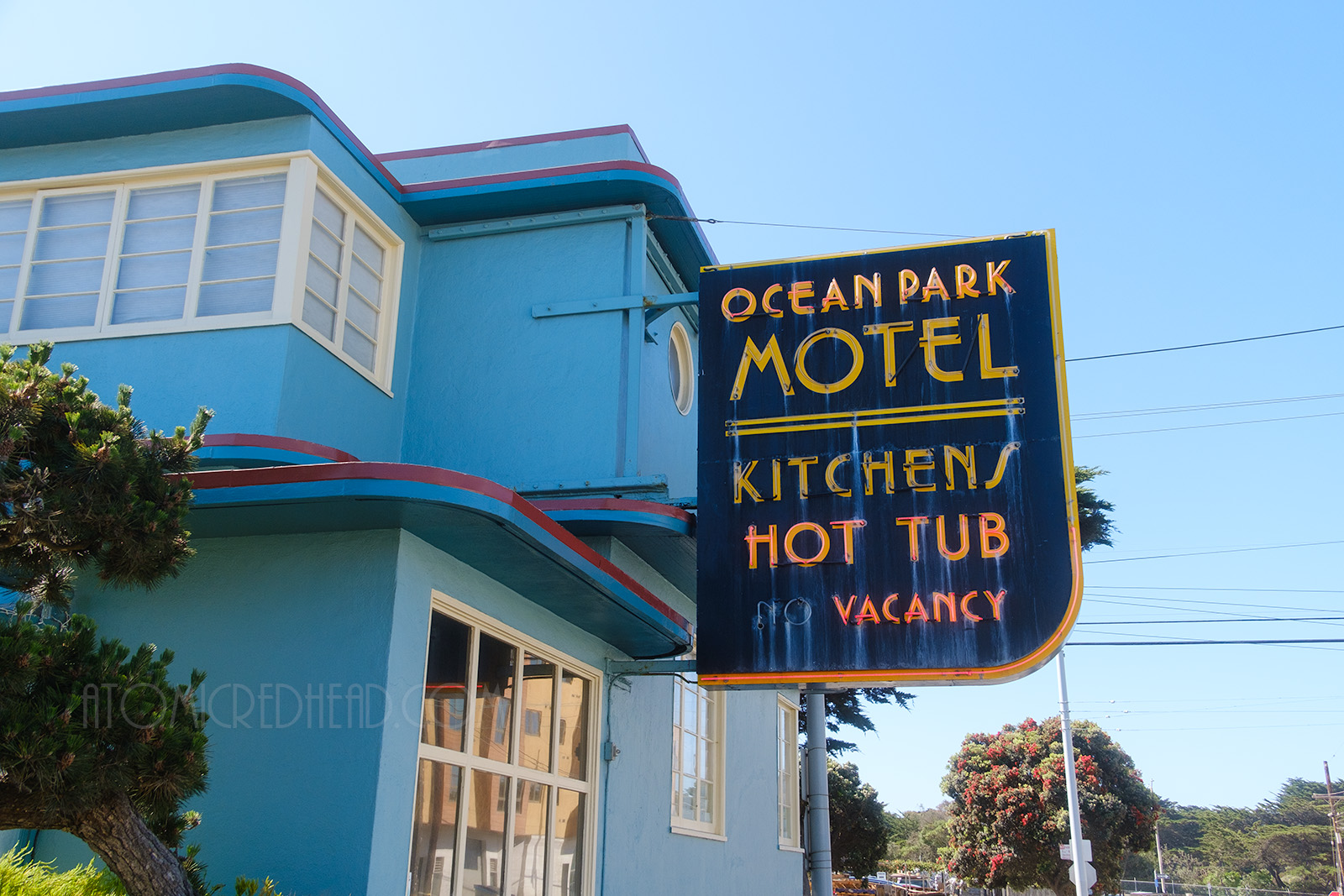 The Ocean Park Motel stands two stories on the corner, with a flat roof that juts out with curved corners, and red trim along the roof edge, the window trim is painted white. The black neon sign reads "Ocean Park Motel Kitchens Hot Tub No Vacancy" in neon.
