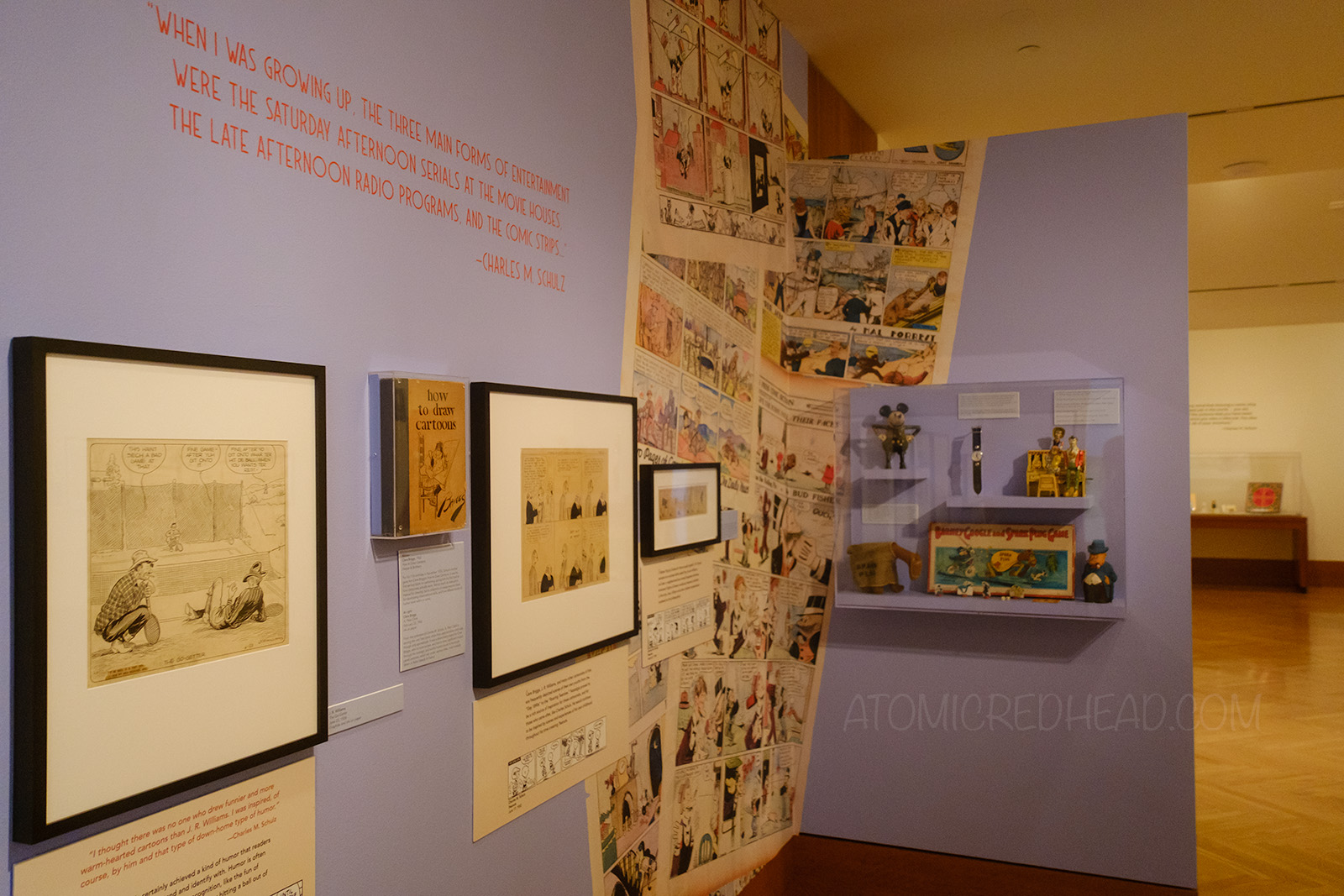 A wall featuring various framed comic strips from the era when Schulz was a child, and a small display case holding vintage toys from the comics.