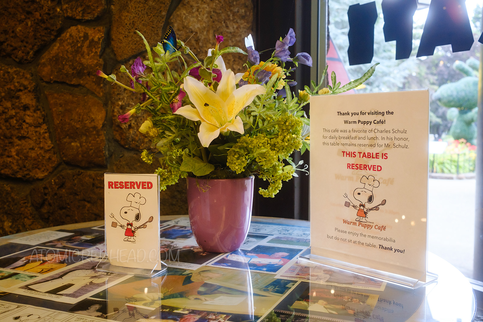 A cafe table featuring a floral arrangement and photos under glass. A sign reads "Reserved" and another reads "That you for visiting the Warm Puppy Cafe! This cafe was a favorite of Charles Schulz for daily breakfast and lunch. In his honor, this table remains reserved for Mr. Schulz. This table is Reserved. Please enjoy the memorabilia but do not sit at the table. Thank you!"