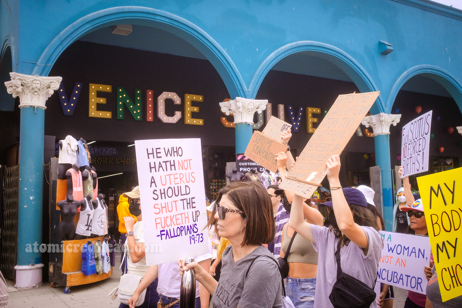 Protestors walk by a blue columned building with large letters reading "Venice" some of the signs read "He who hath not a uterus should shut the fucketh up" another reads "I'm a woman not an incubator" another reads "Fuck SCOTUS No Forced Births"