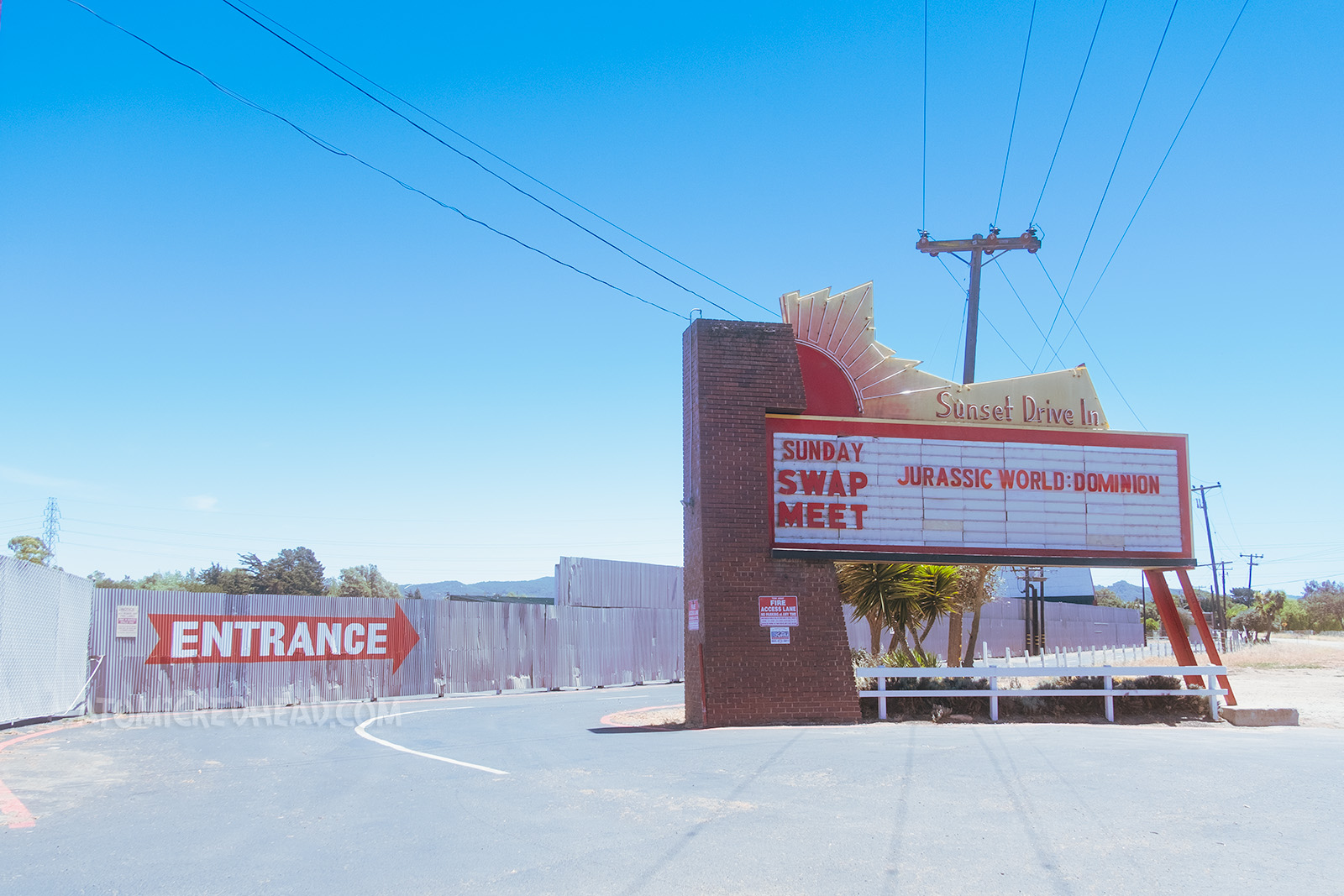 Marquee and entrance for the Sunset Drive-In, which features a neon sun setting, with neon letters reading "Sunset Drive-In" on the marquee it reads "Sunday Swap Meet Jurassic World: Dominion" in red letters. A corrugated metal fence behind the marquee has a large red arrow with white letters inside that read "Entrance" 
