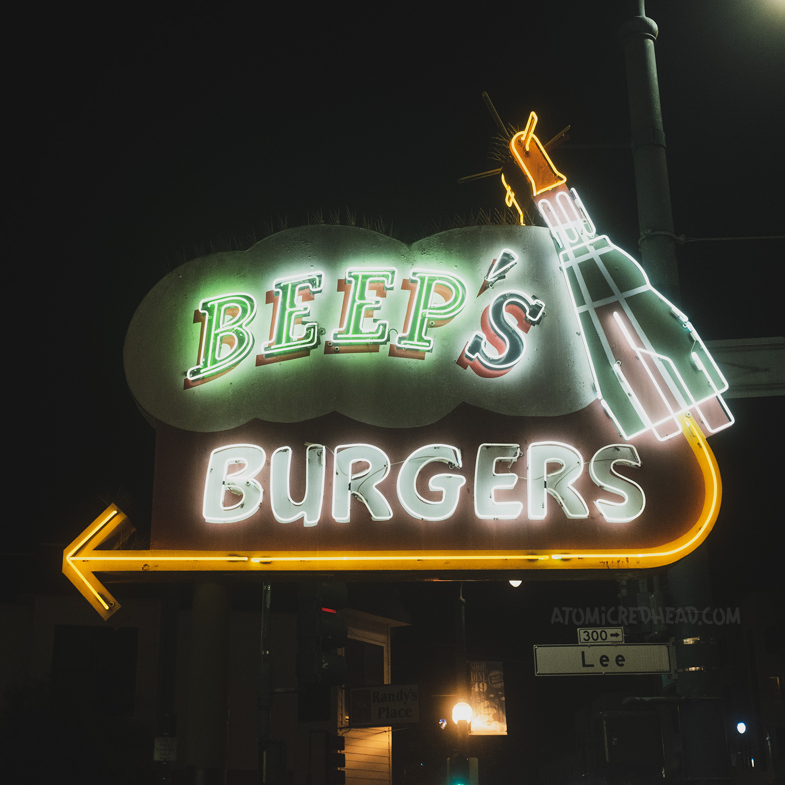 A neon space capsule rises on the right side of the Beep's Burgers sign, with "Beep's Burgers" spelled out in neon. A gold neon arrow points to the left. 