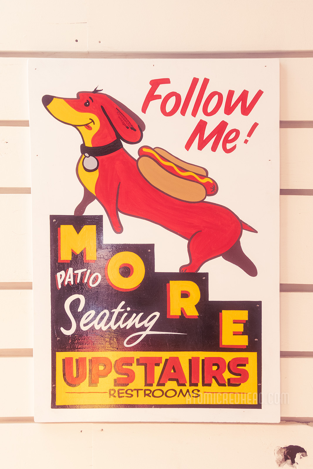 A small painted sign features a dachshund with a hot dog on his back going up a set of stairs, text reads "Follow Me! More patio seating upstairs, restrooms"