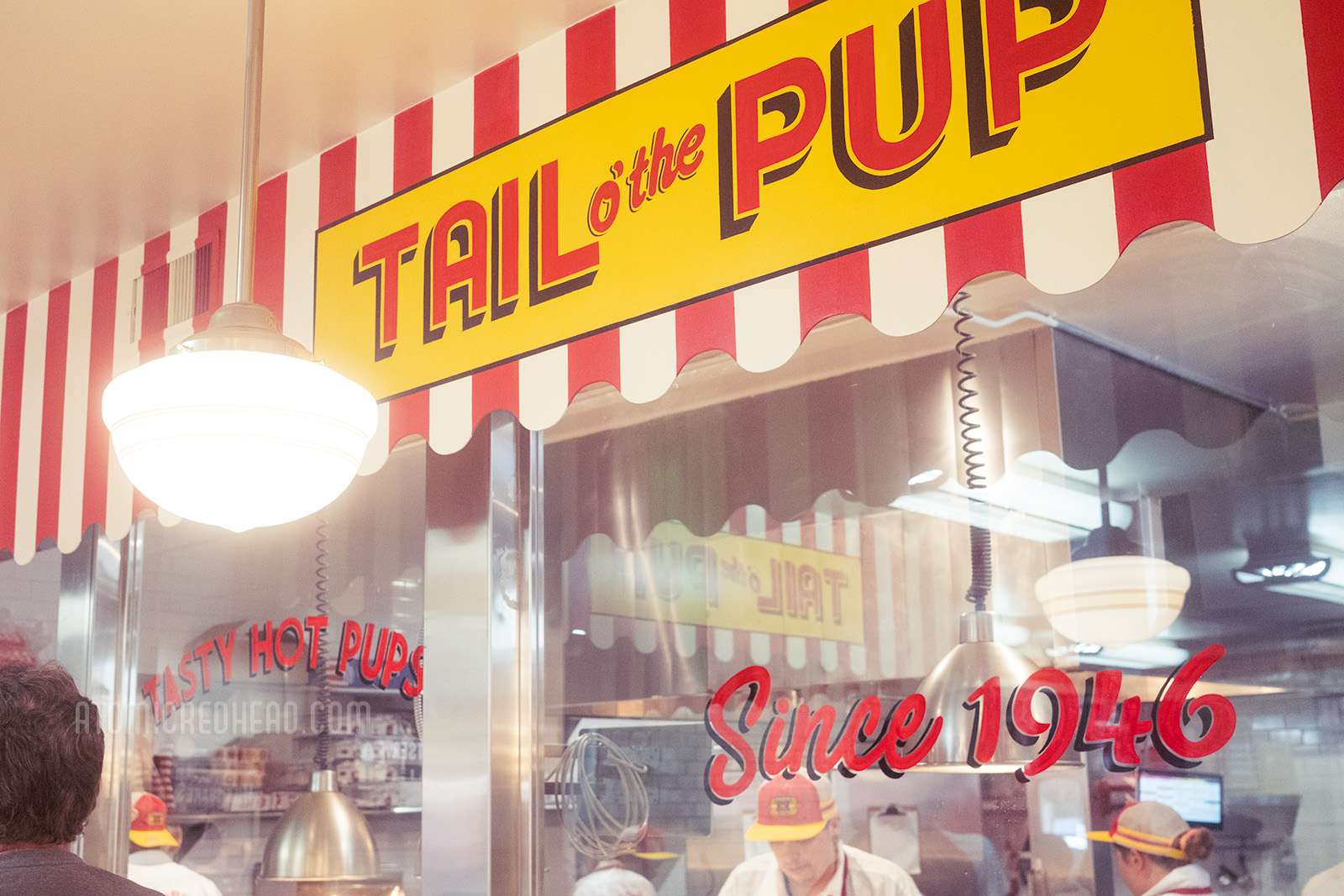 Behind a window various employees dish up orders. Above a red and white stripe sign reading "Tail o' the Pup" and across the window reads "Since 1946"
