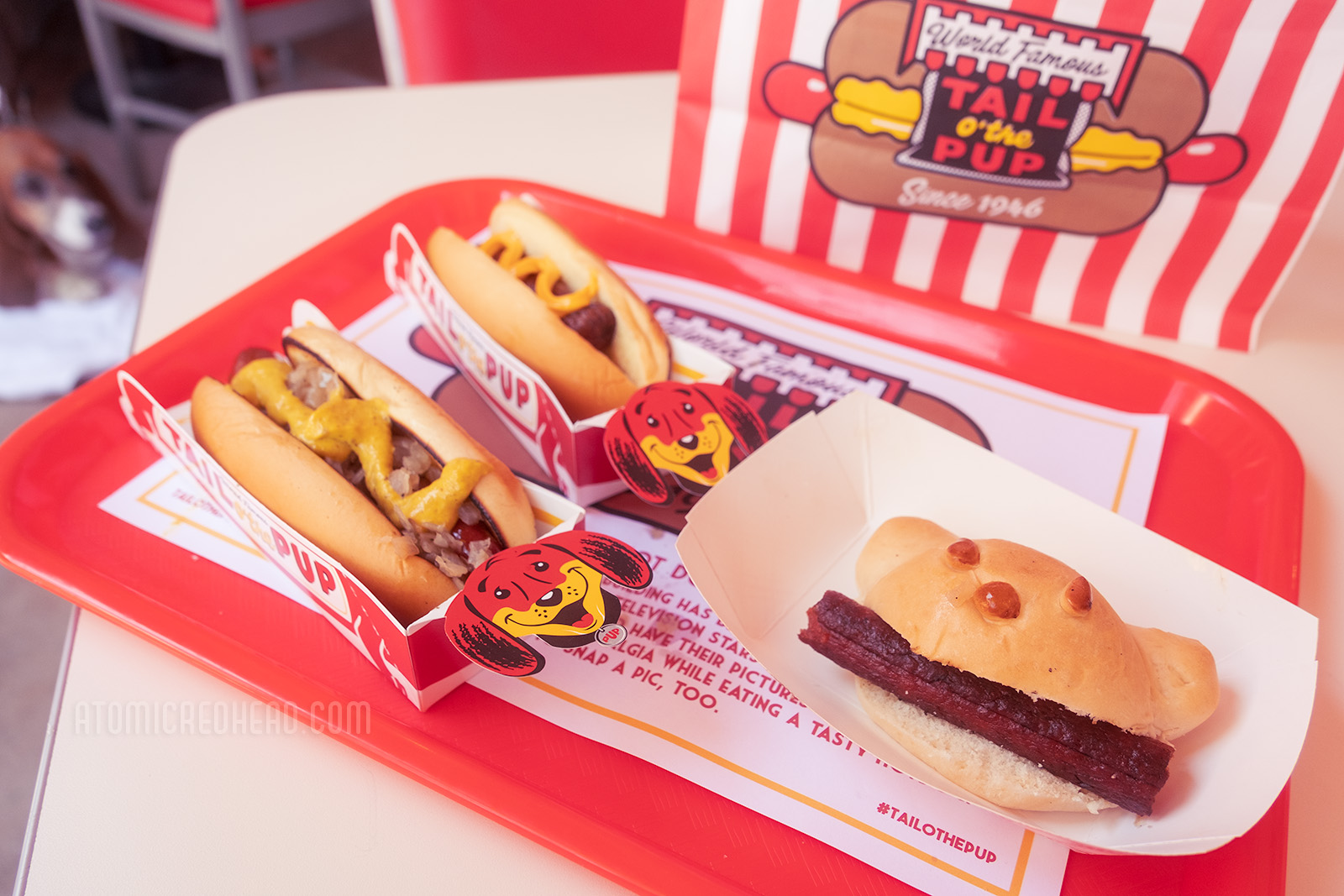 On a red tray sit two cardboard containers featuring dog faces hold two hot dogs. In another cardboard boat sits a bun shaped like a dog, with a hot dog in its mouth.