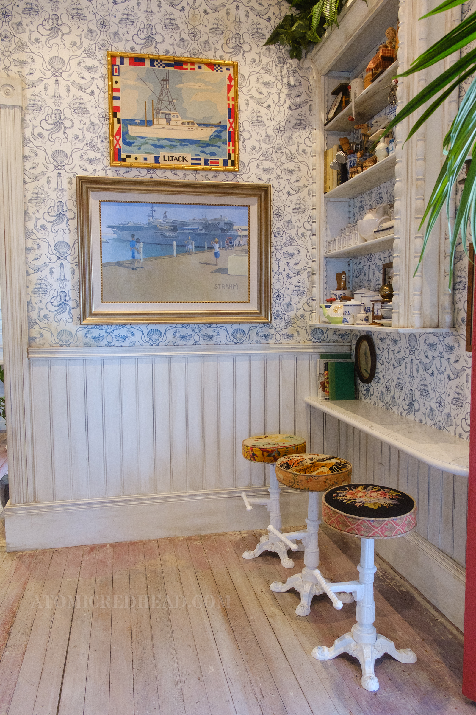 A small counter seats three, the walls are covered in white and blue wallpaper that is of a nautical theme, a painting of a beach hangs on the wall.