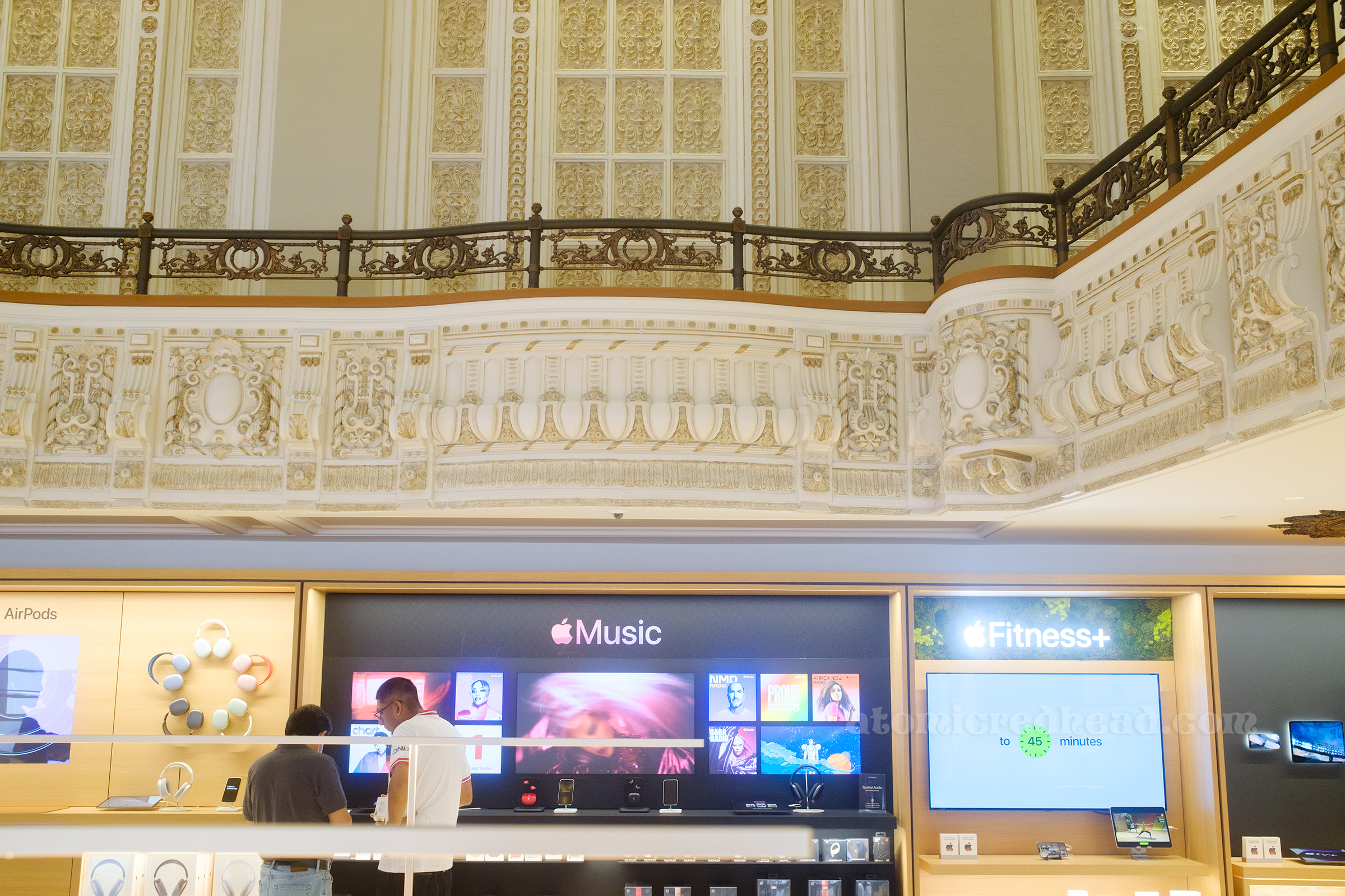 Interior of store, on the ground level clean lines display screens and Apple products, above a highly ornate balcony painted in white and gold.