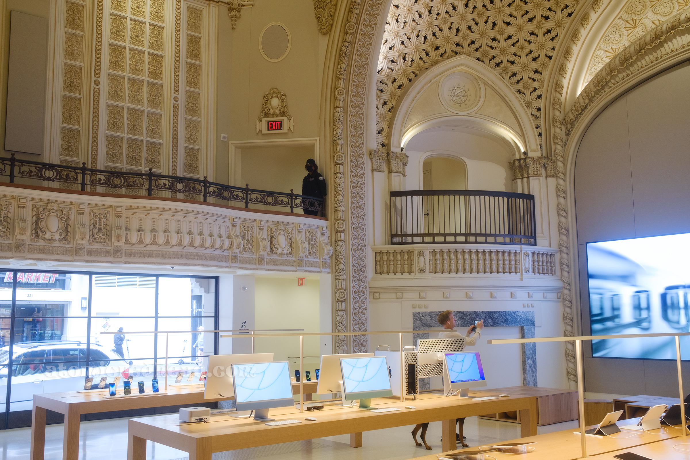 Overview of part of the sales floor, which includes light colored wood tables in the middle with various Apple products such as iMacs and laptops on display. In the back is an arched balcony framed in white and gold.