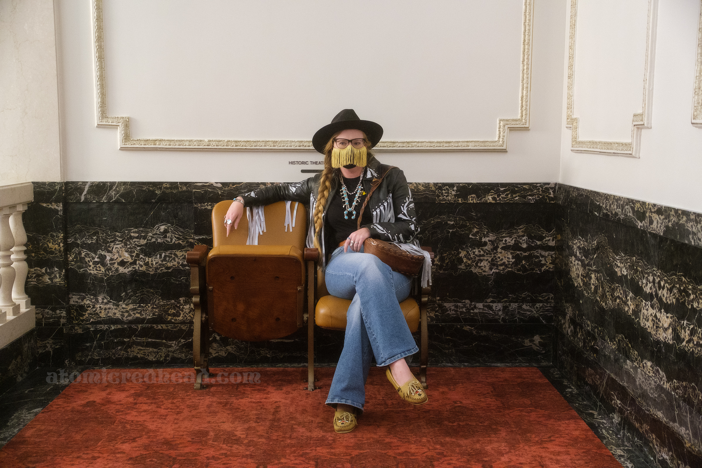 Myself, seated in one of the original theatre chairs, wearing a black cowboy hat, black and silver fringe leather jacket, black shirt, and blue jeans.
