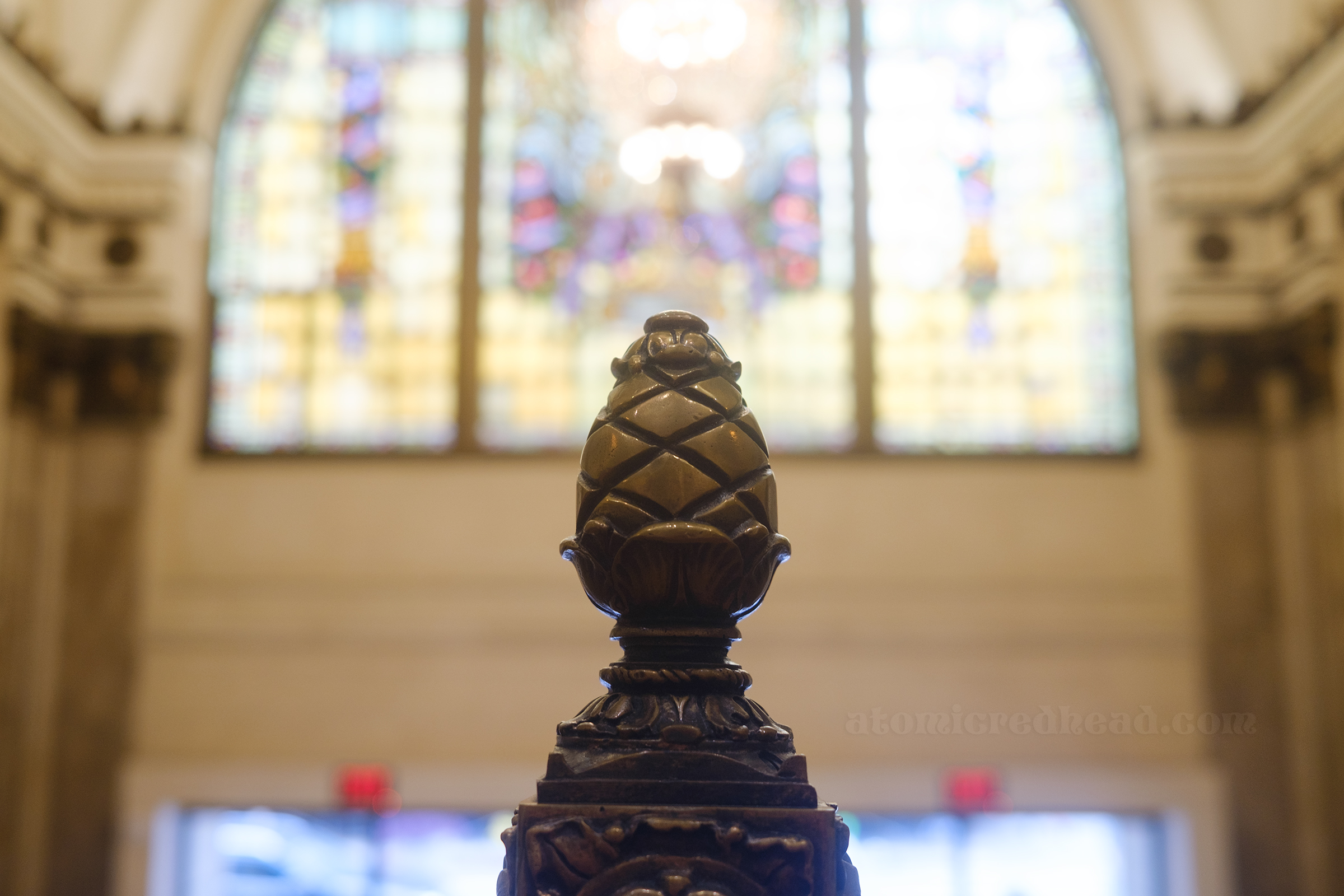 Close-up of a brass pinecone that sits atop the railing, in the distance is a massive stained glass window.