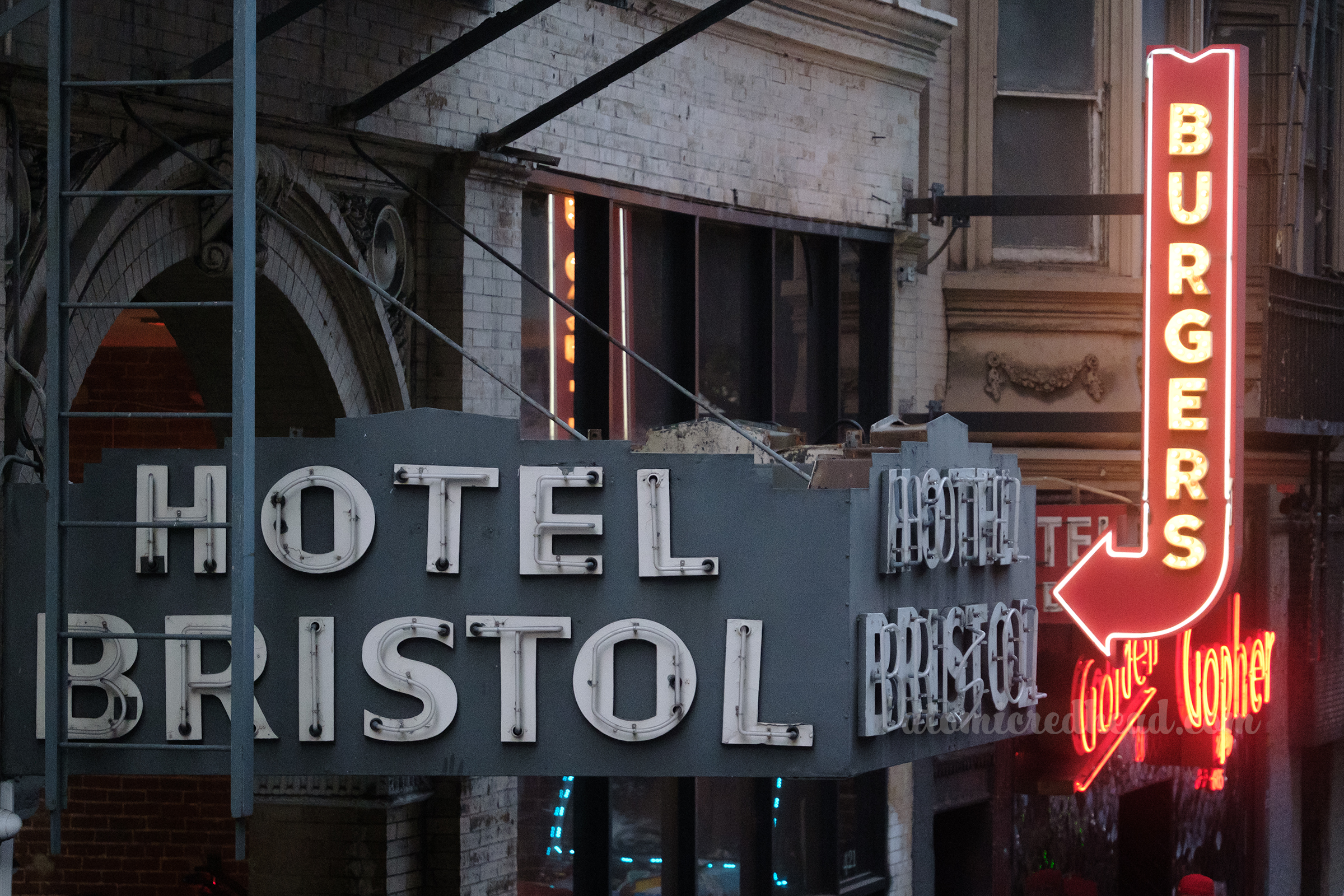 A three sided neon sign juts out above a doorway, and reads "Hotel Bristol" on each side. A red arrow, edged in neon reads "Burgers" beyond it.